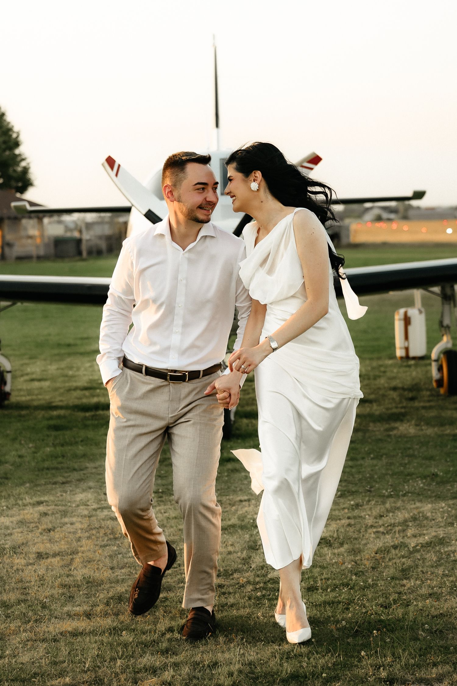 A smiling couple in wedding attire walks hand-in-hand on a grassy airfield with a small plane in the background.