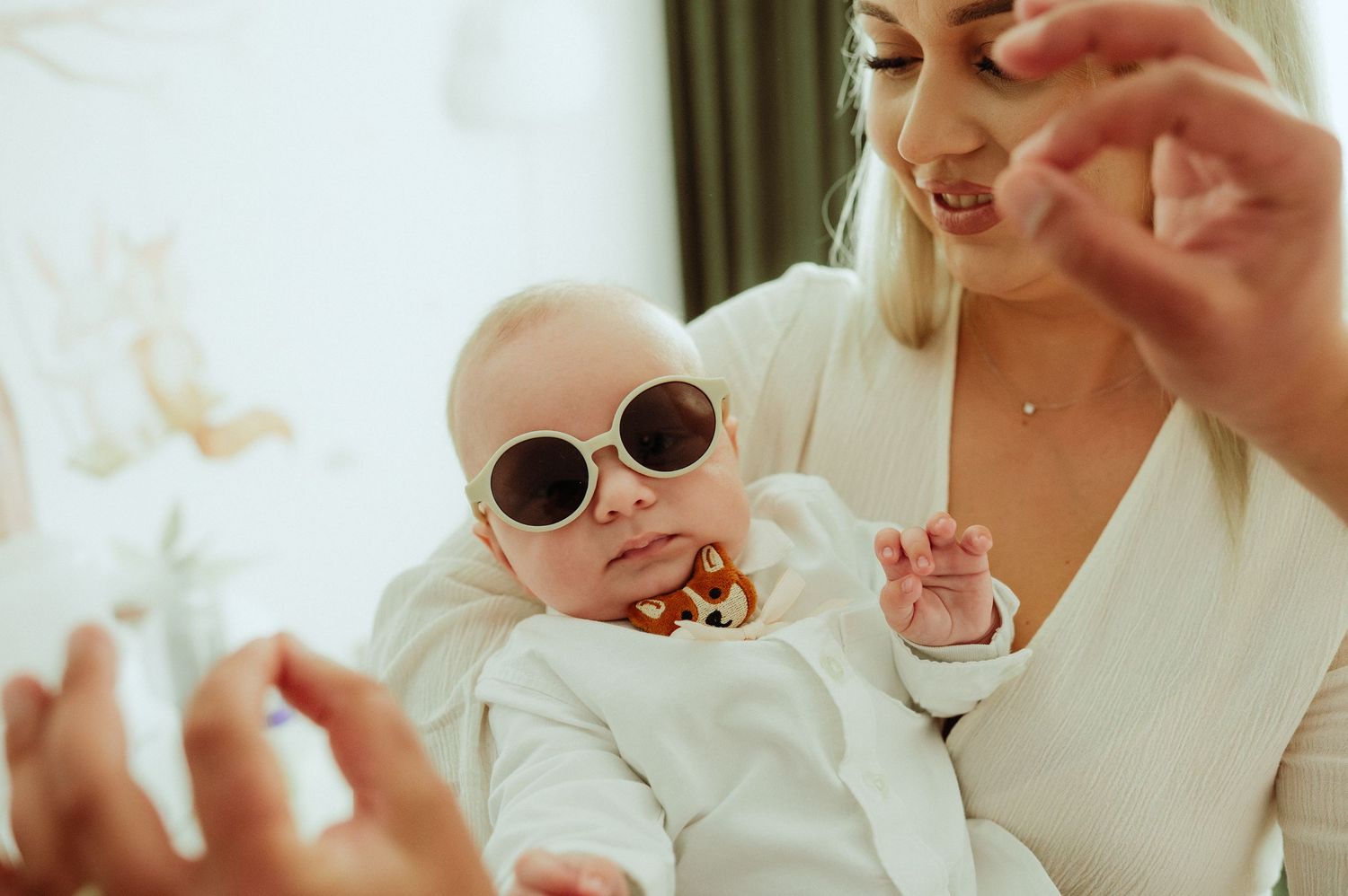Adorable baby wearing round sunglasses held by smiling blonde mother in white outfit.