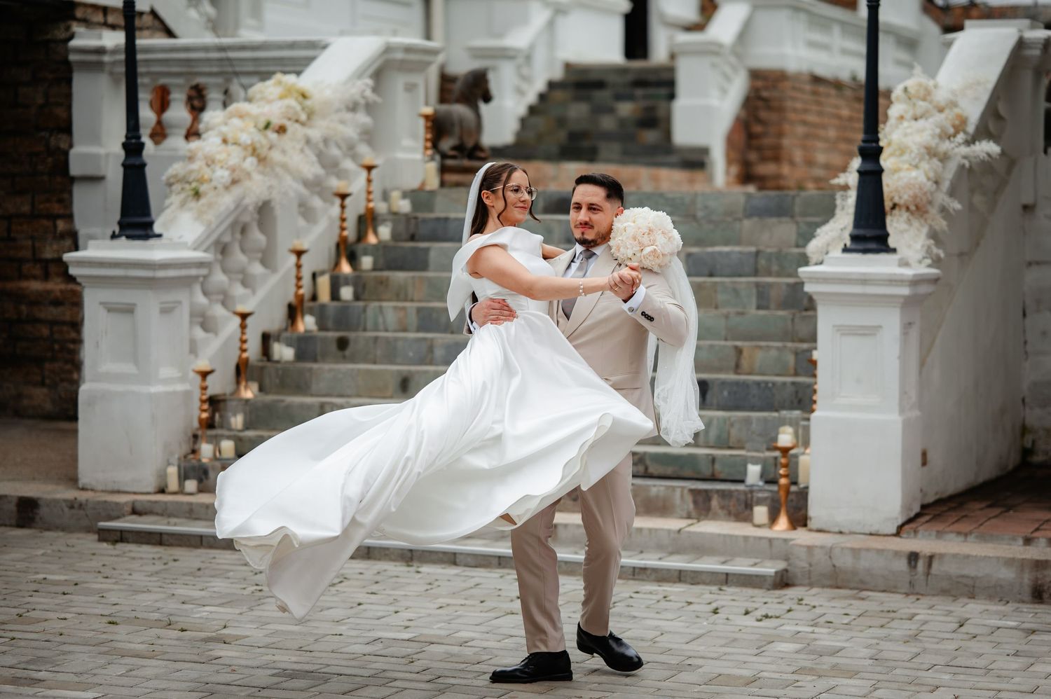 Groom lifts bride in white flowing gown as she laughs joyfully on cobblestone courtyard at elegant wedding venue.