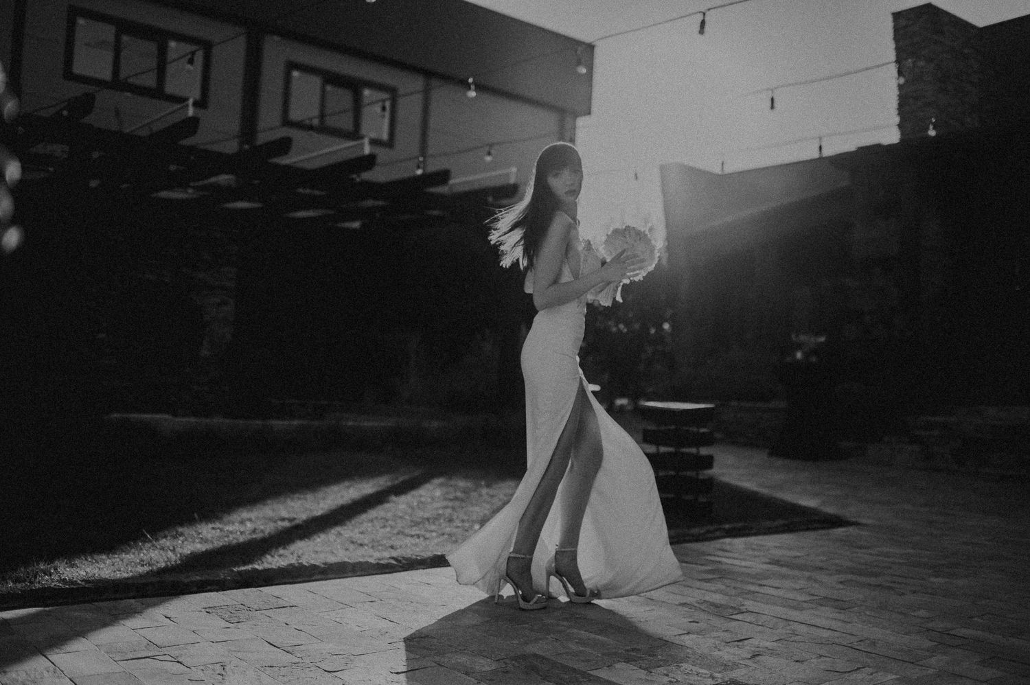 Black and white artistic wedding photo of bride in elegant gown twirling with bouquet in sunlight.