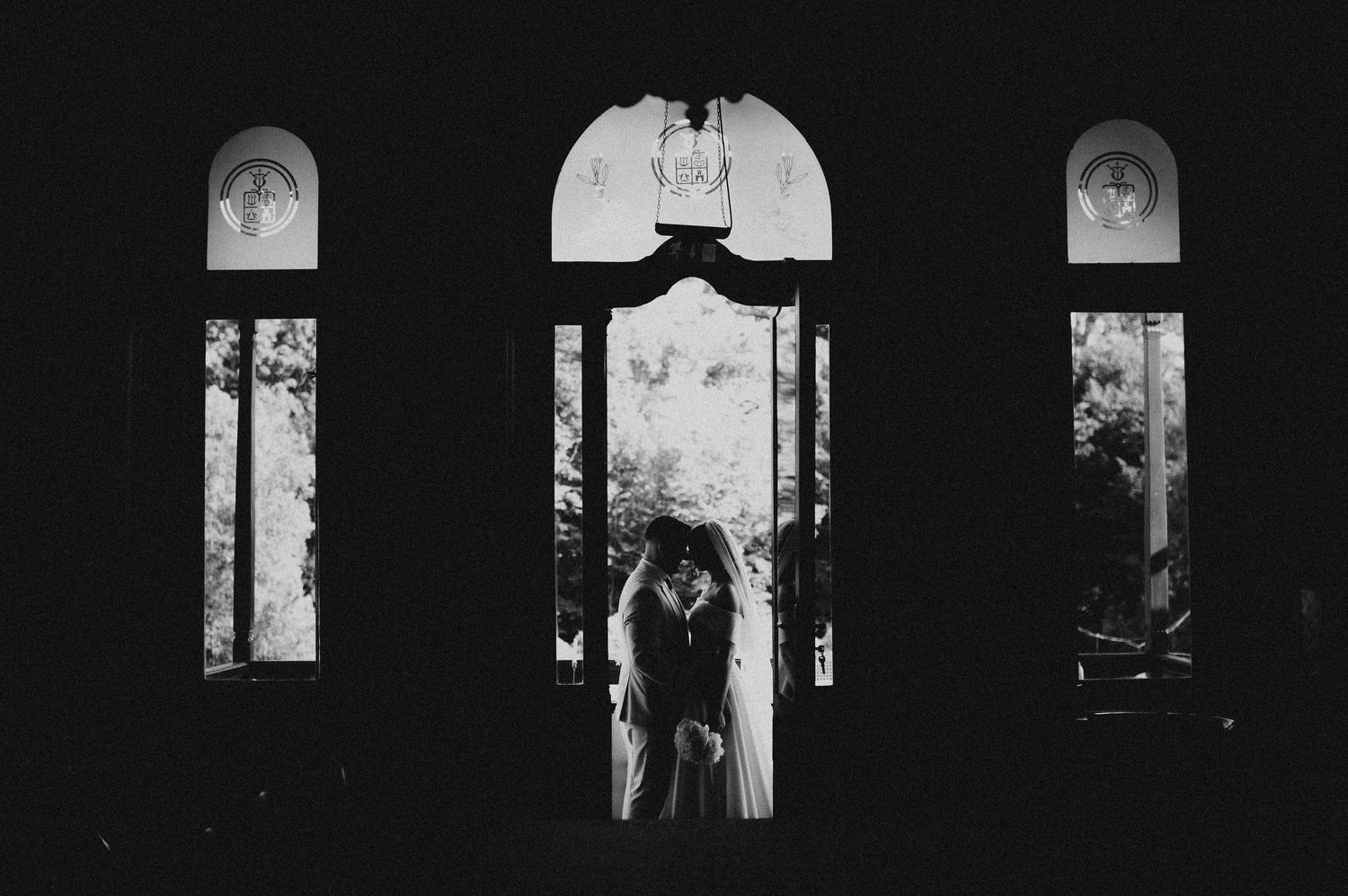 Dramatic black and white silhouette of couple embracing in arched doorway with garden light streaming through windows.