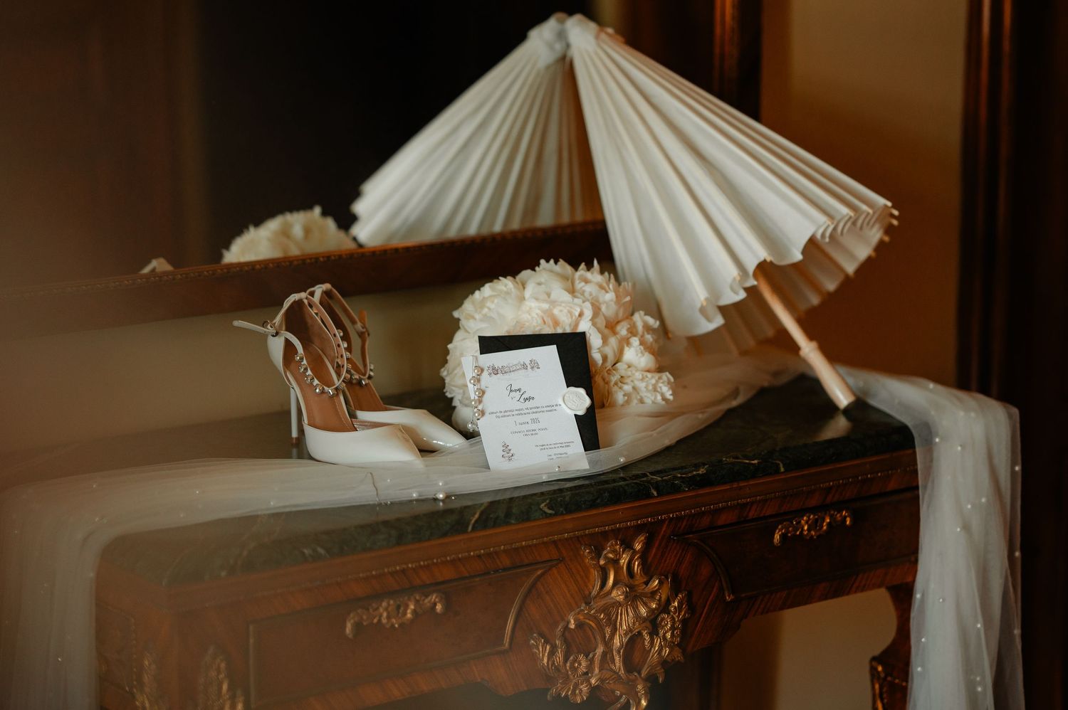 Wedding flat lay with white parasol, bridal heels, invitation, and floral details on antique dresser.