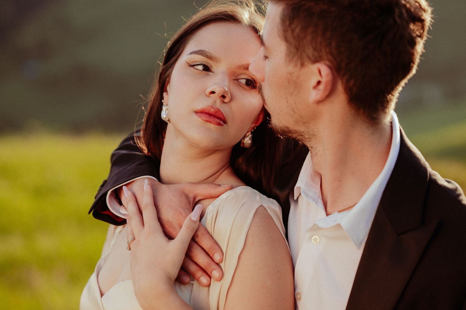 Groom in black suit tenderly kisses bride on cheek while embracing her in a sunlit field.