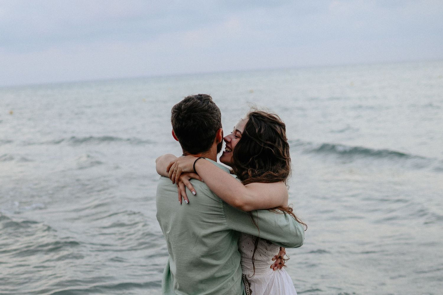 Loving couple embracing and sharing a tender moment facing the calm open sea on an overcast day.