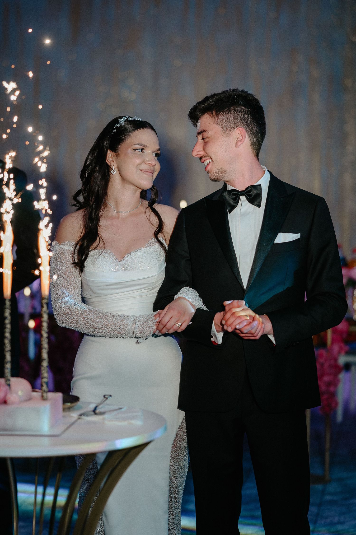 Smiling bride in beaded white gown and groom in black tuxedo stand together near wedding cake.