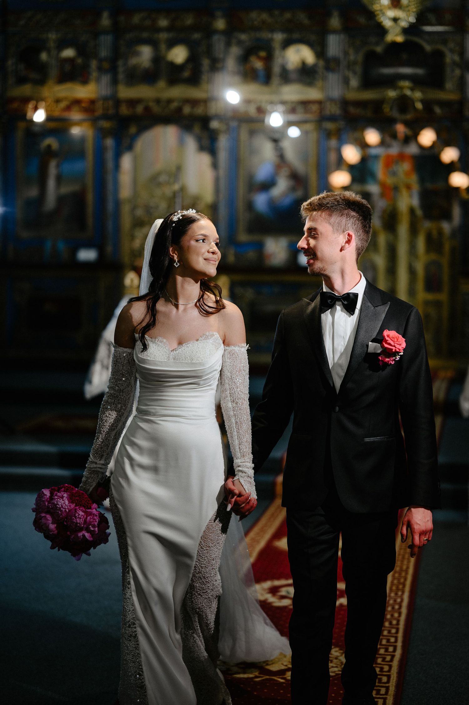 Bride and groom walk hand in hand through ornate Orthodox church interior after wedding ceremony.