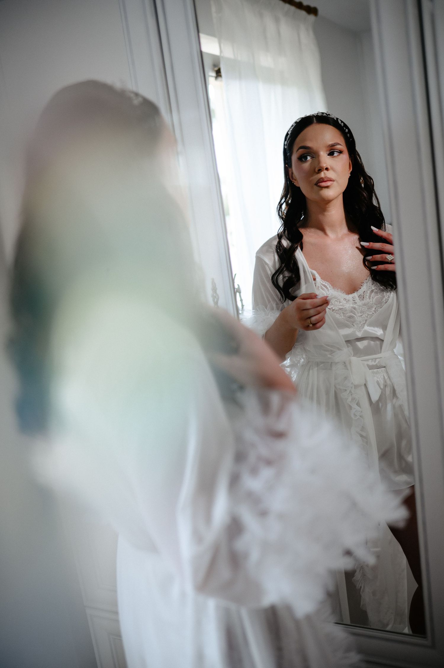 Bride in white lace robe admiring herself in full-length mirror during bridal morning preparations.