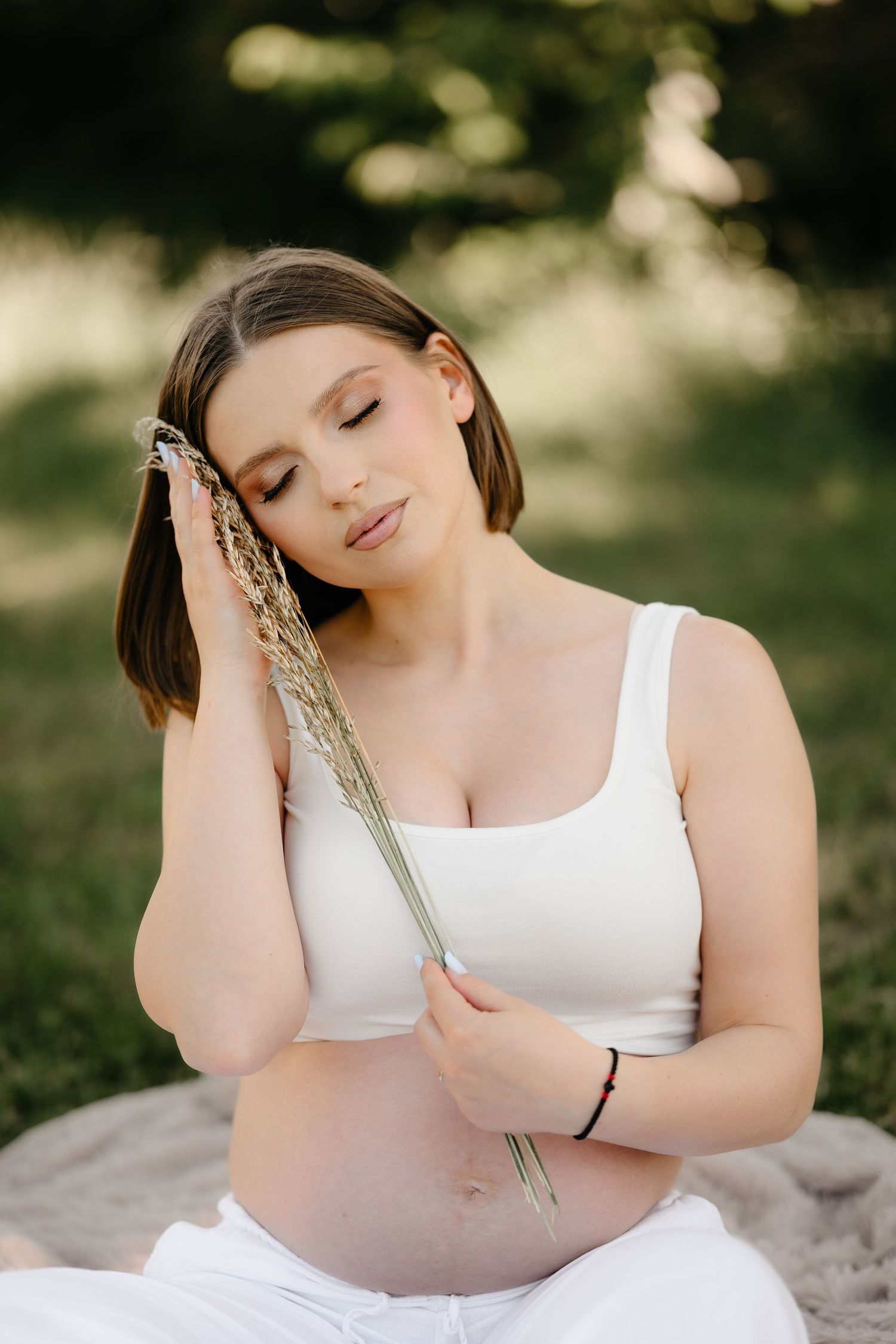 Serene pregnant woman in white sitting outdoors, eyes closed, holding dried wheat grass.