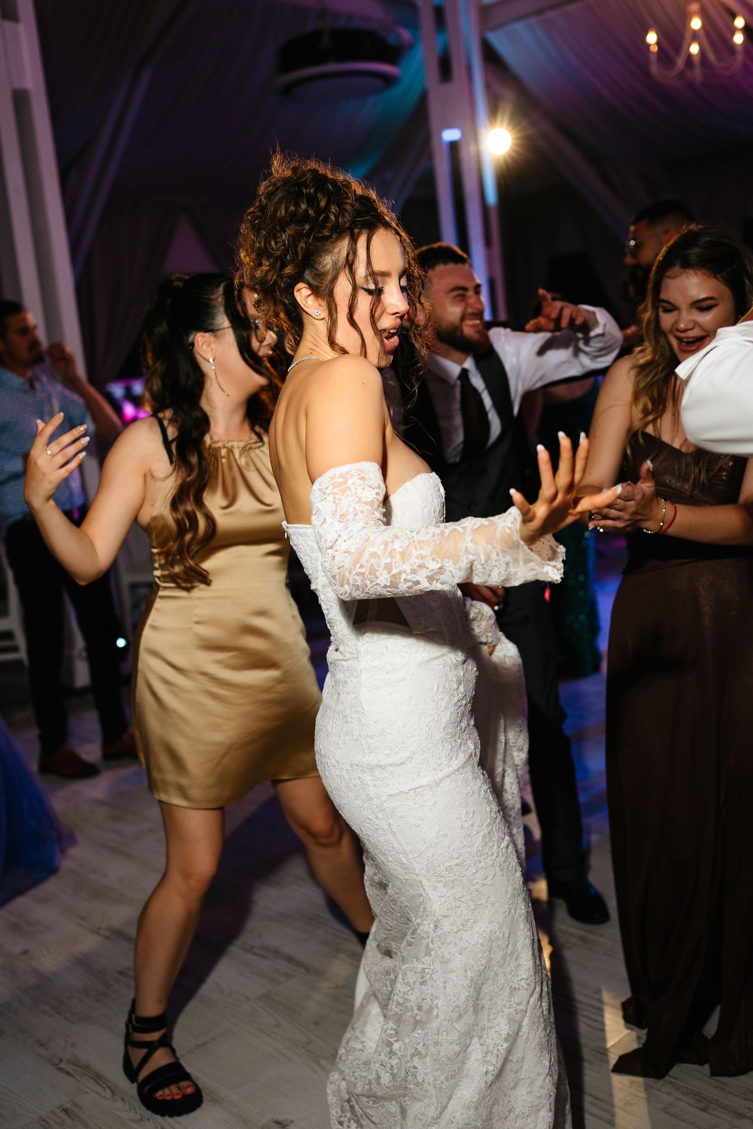Bride in white lace gown dancing joyfully with guests at a wedding reception party.