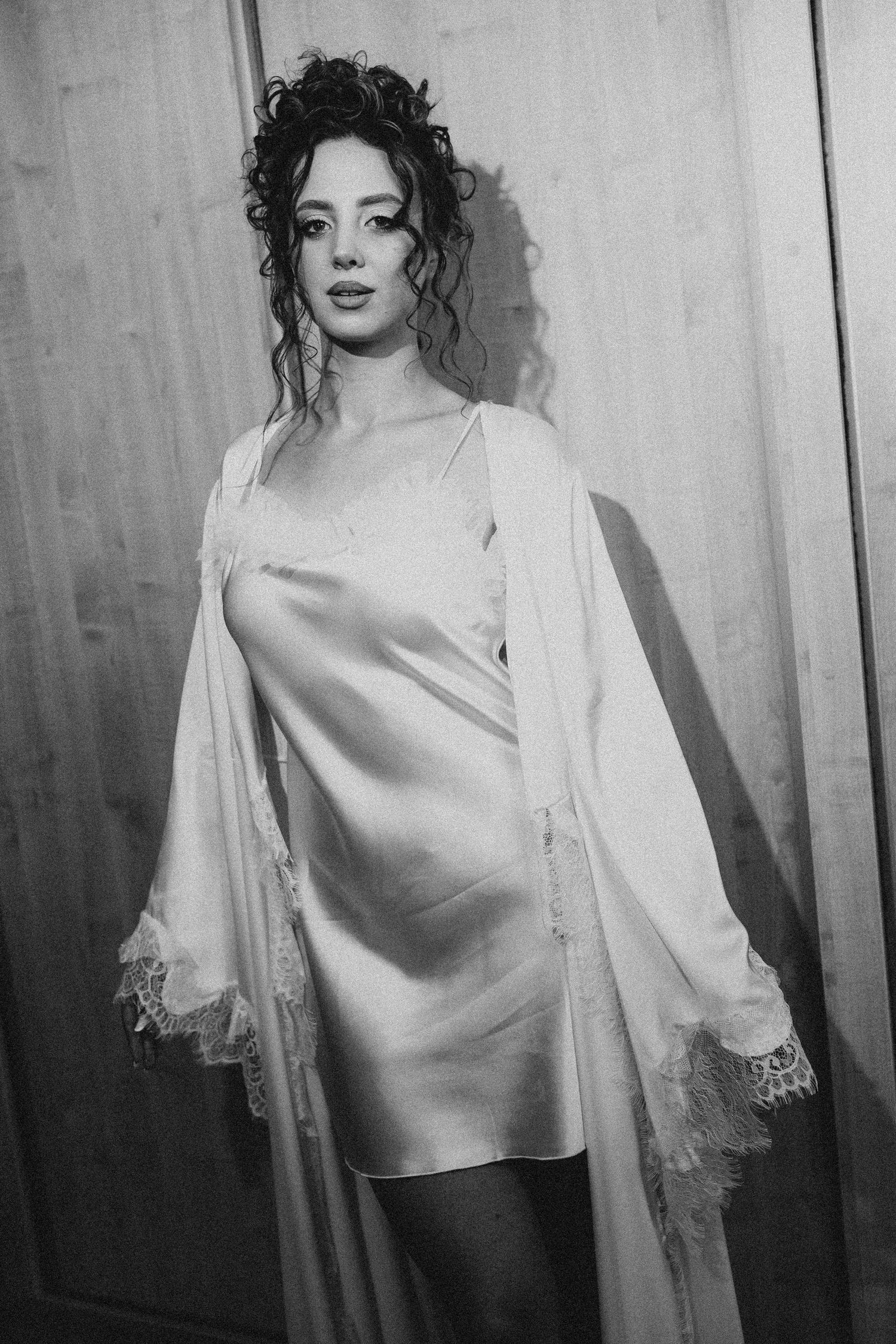 Black and white photo of a woman in a satin slip dress and lace robe, posing elegantly against wooden doors.