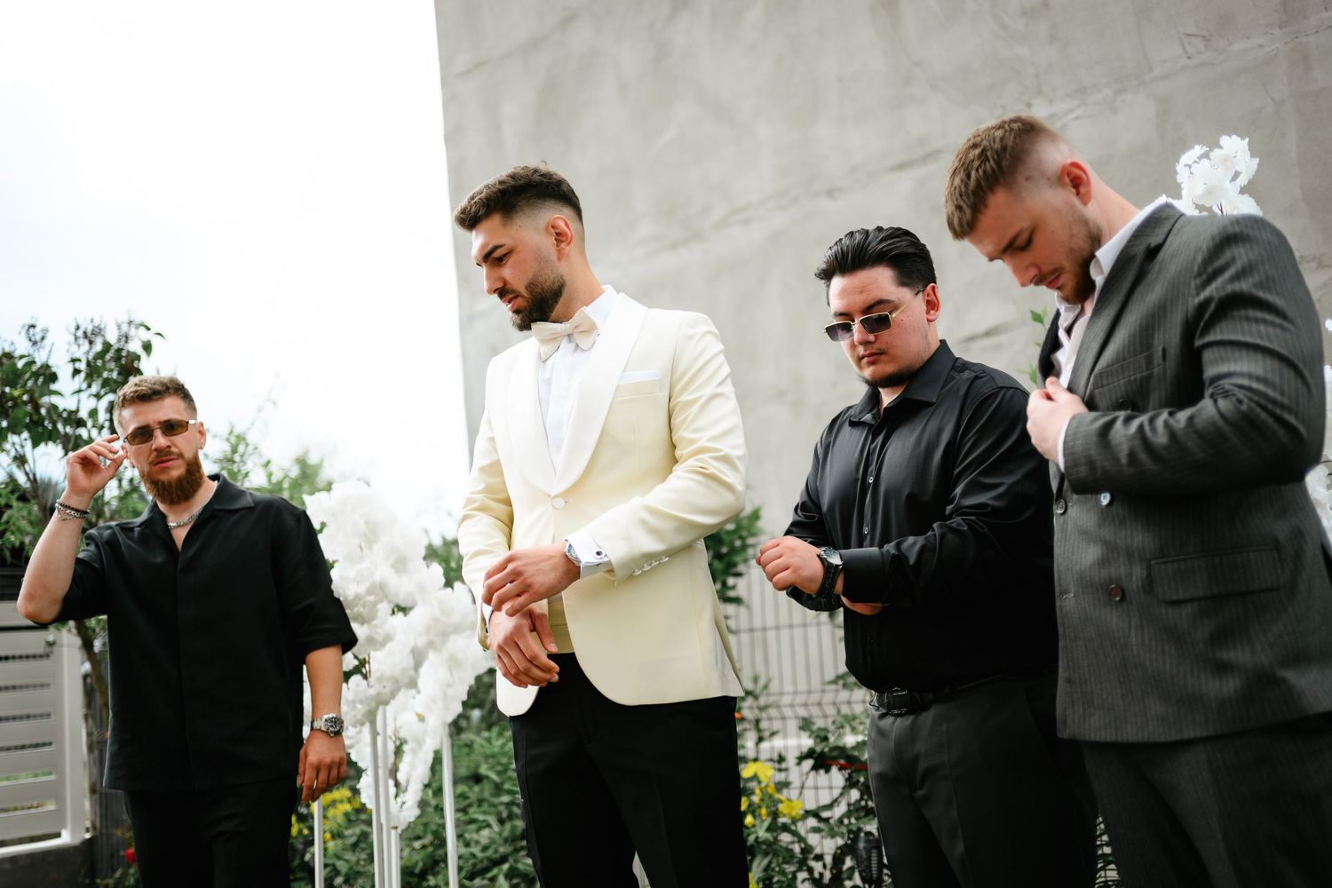 Groom in white tuxedo stands with groomsmen in black suits outdoors before wedding ceremony.