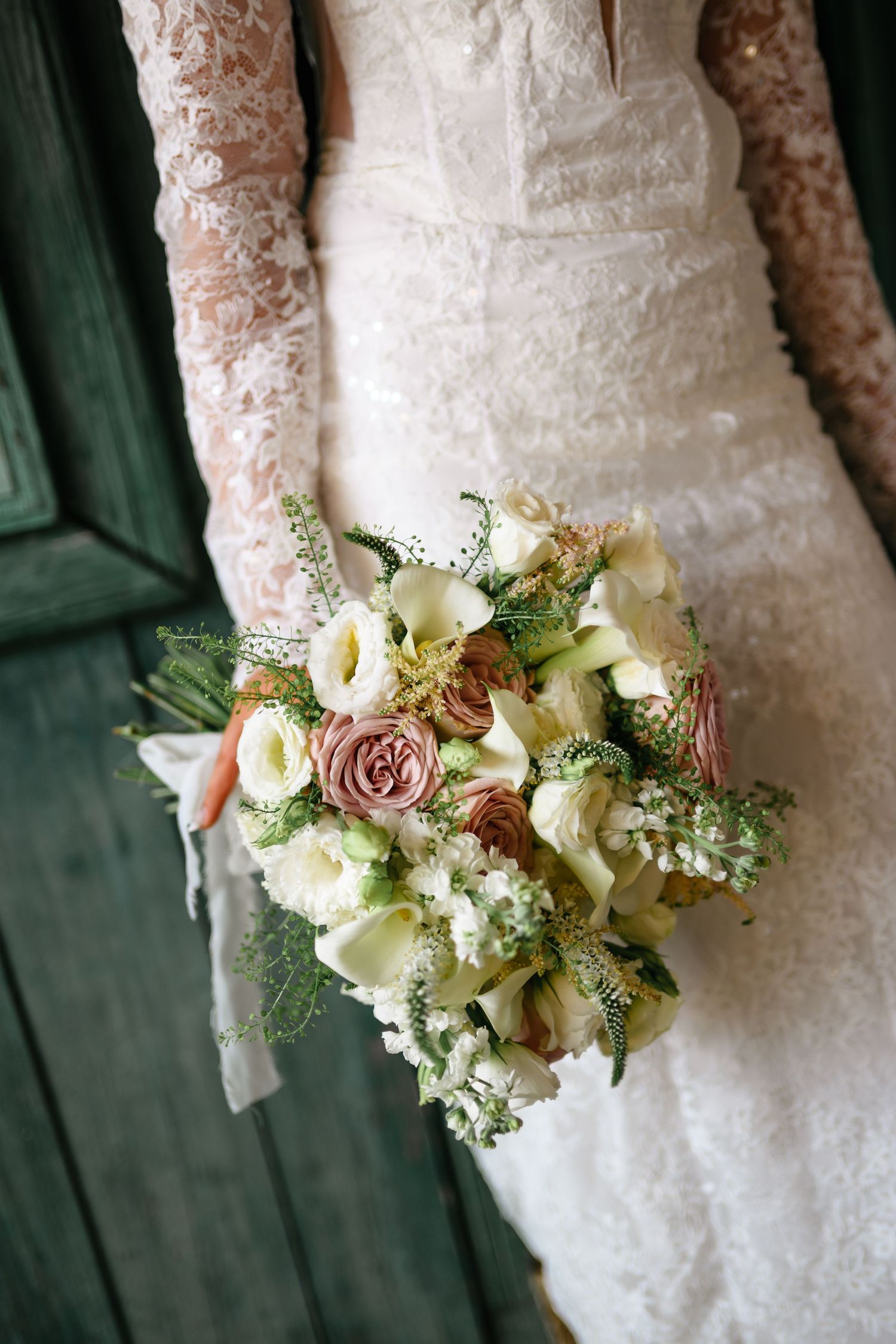Bride in lace wedding dress holding a romantic bouquet of pink roses, white calla lilies, and greenery by a green door.
