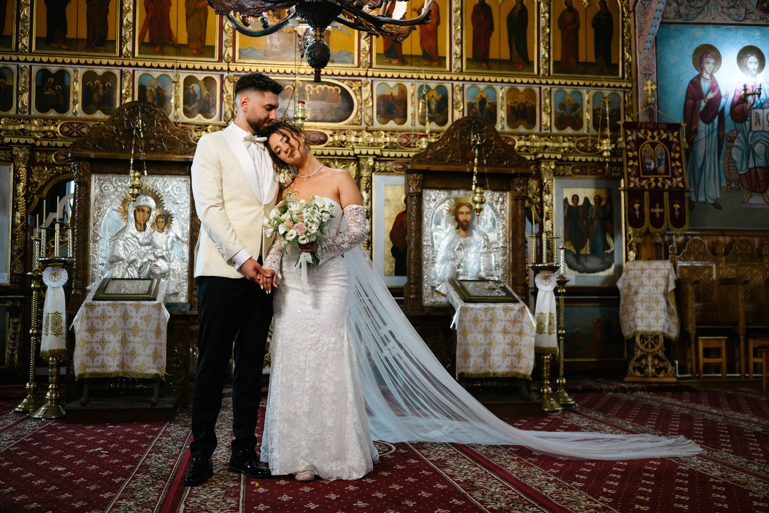 Bride and groom posing together inside an ornate Orthodox church with golden iconostasis during their wedding ceremony.