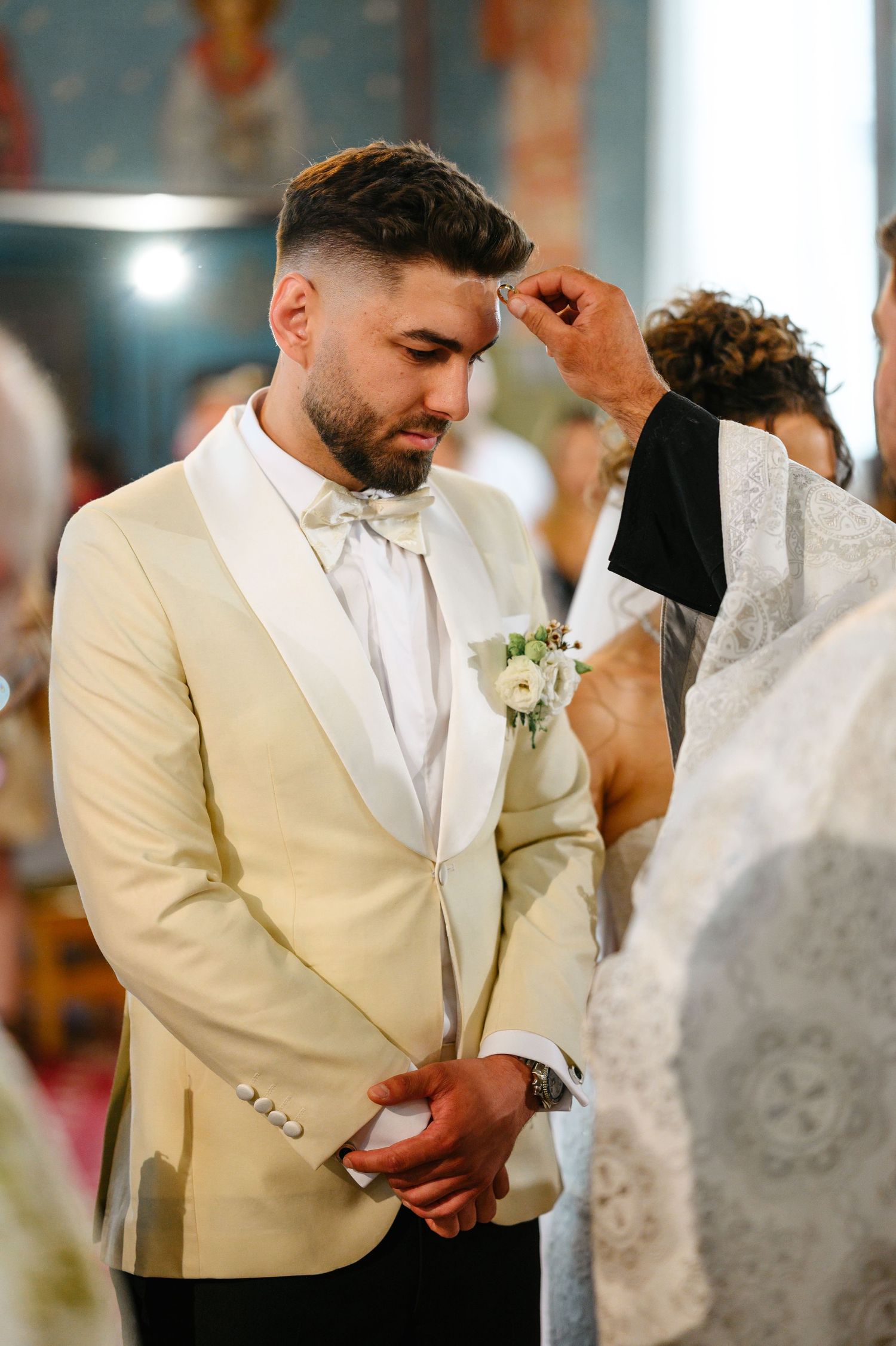 Groom in ivory tuxedo receives blessing from priest during elegant church wedding ceremony.