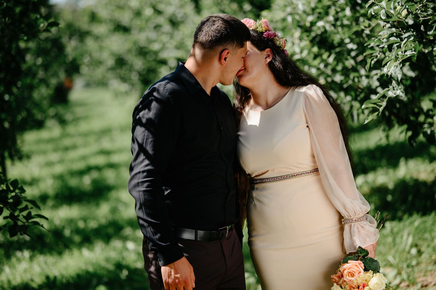 Bride and groom sharing a romantic kiss in a lush green orchard, bride holding a colorful floral bouquet.