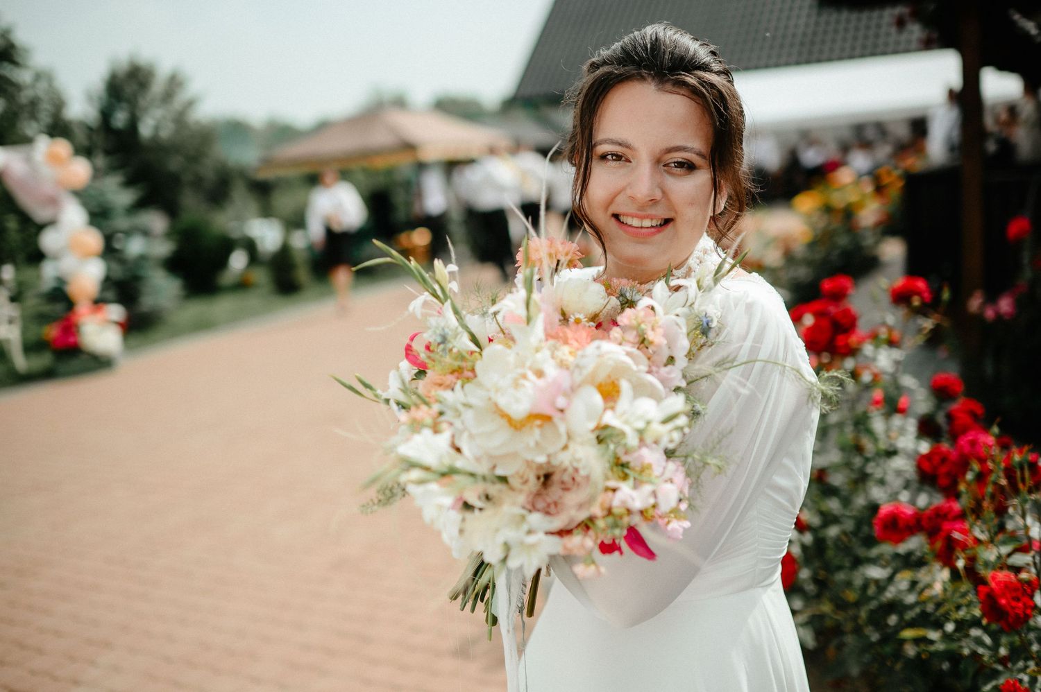Smiling bride in white dress holds floral bouquet near rose garden outdoor venue.