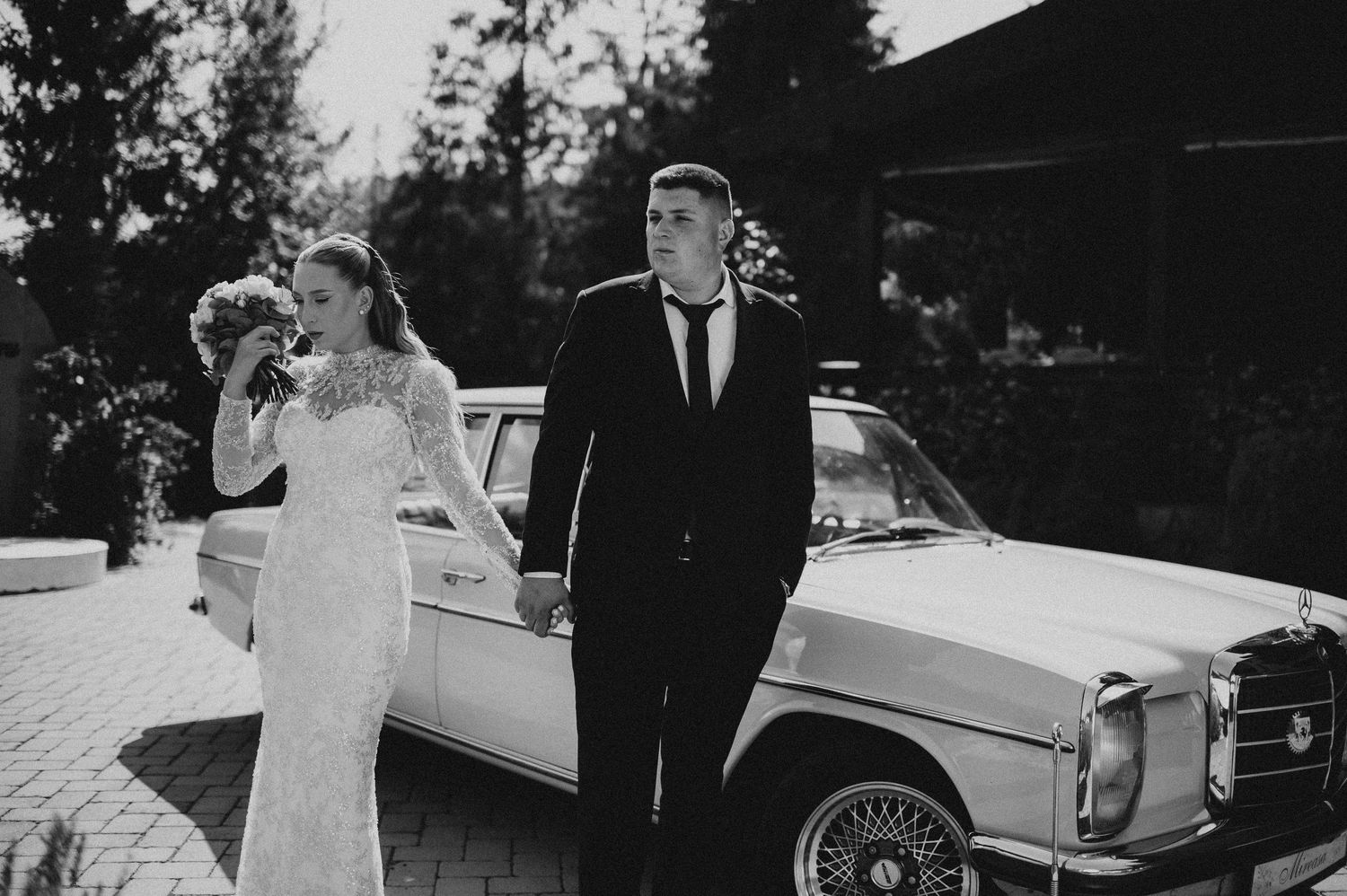 Black and white photo of a bride and groom standing beside a classic Mercedes-Benz car on their wedding day.