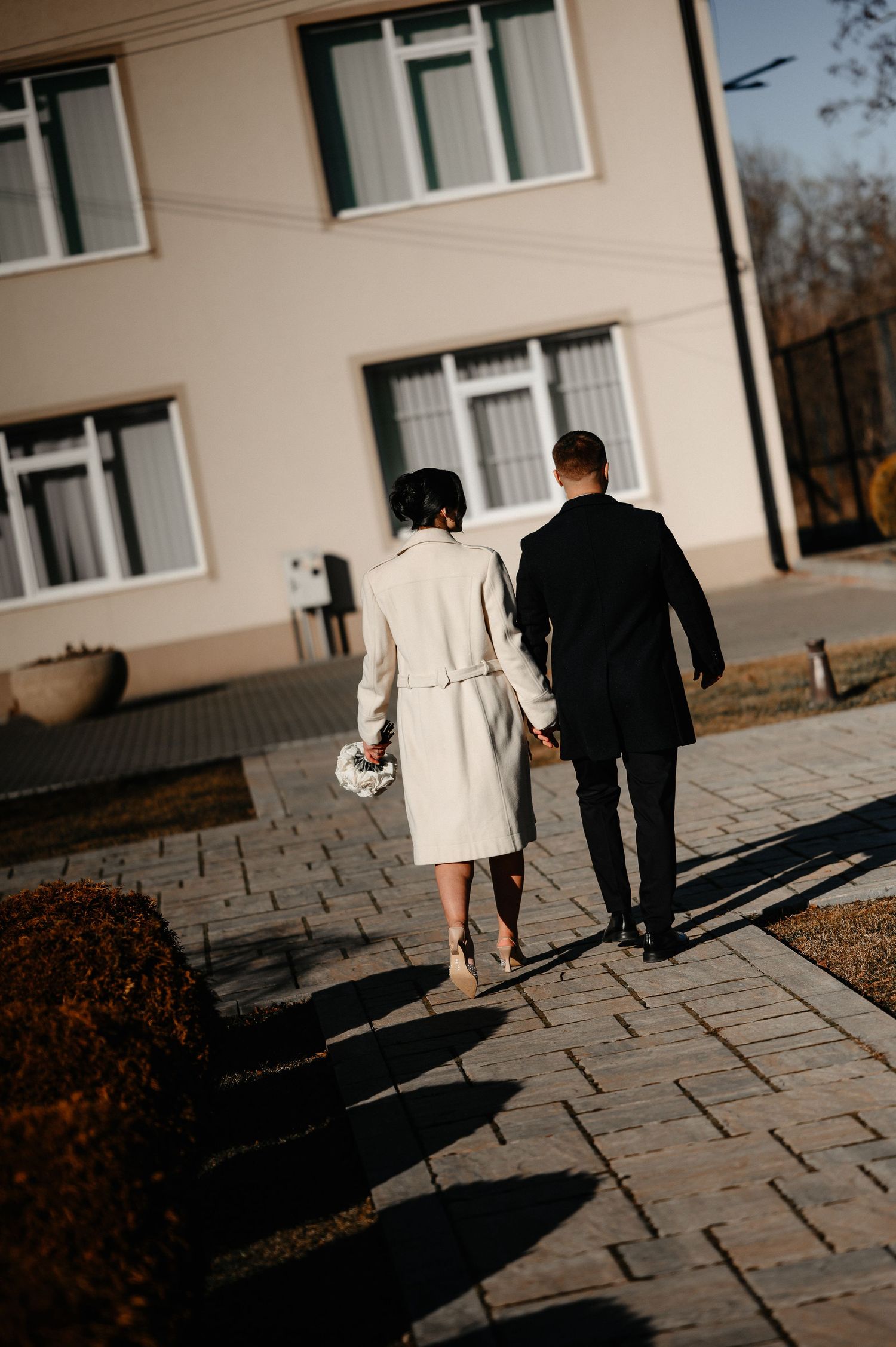 Bride and groom walking hand in hand on a sunny paved path toward a building.
