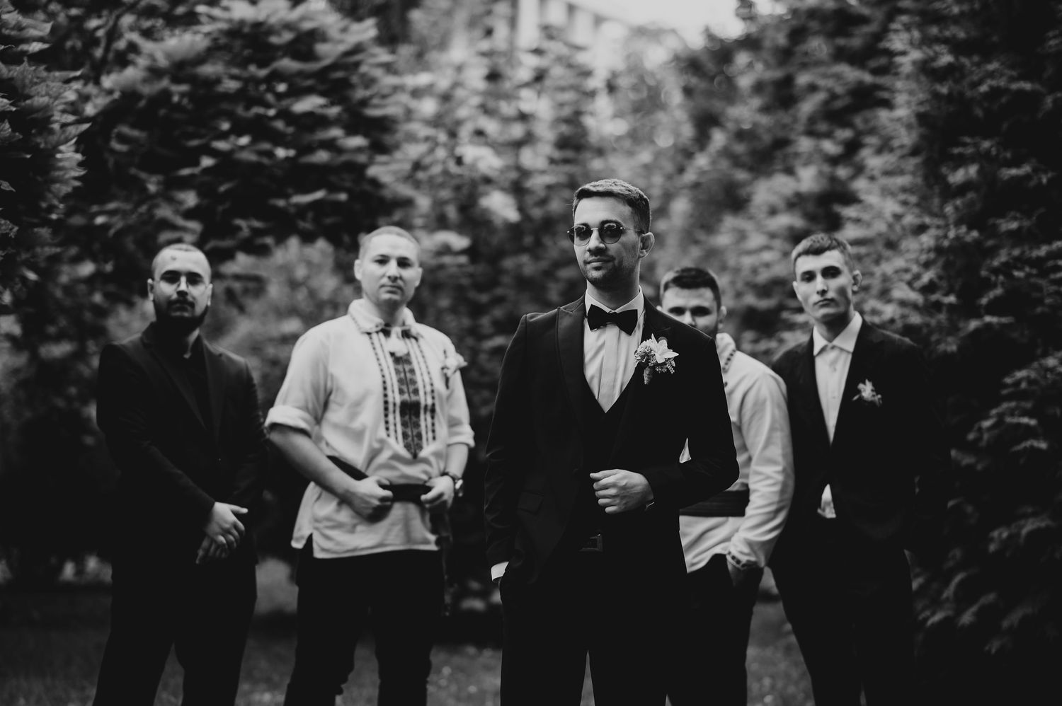 Groom in tuxedo poses with groomsmen in black suits outdoors in black and white photo.