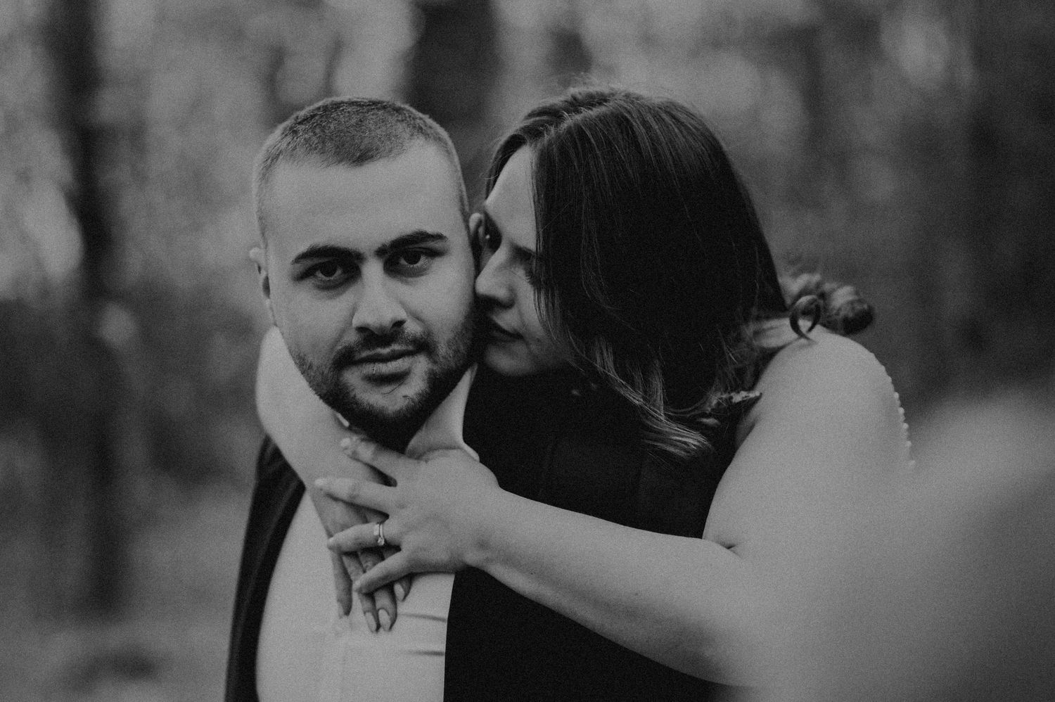 Black and white photo of a couple embracing in a forest, woman hugging man from behind in a romantic outdoor setting.