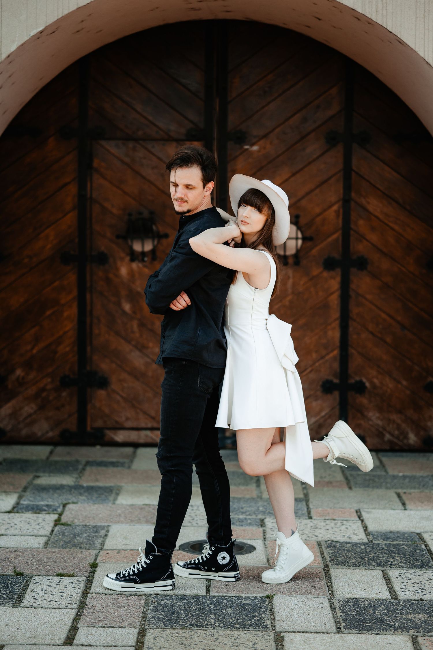 Woman in white dress hugging man in black from behind, posing before a rustic wooden door.