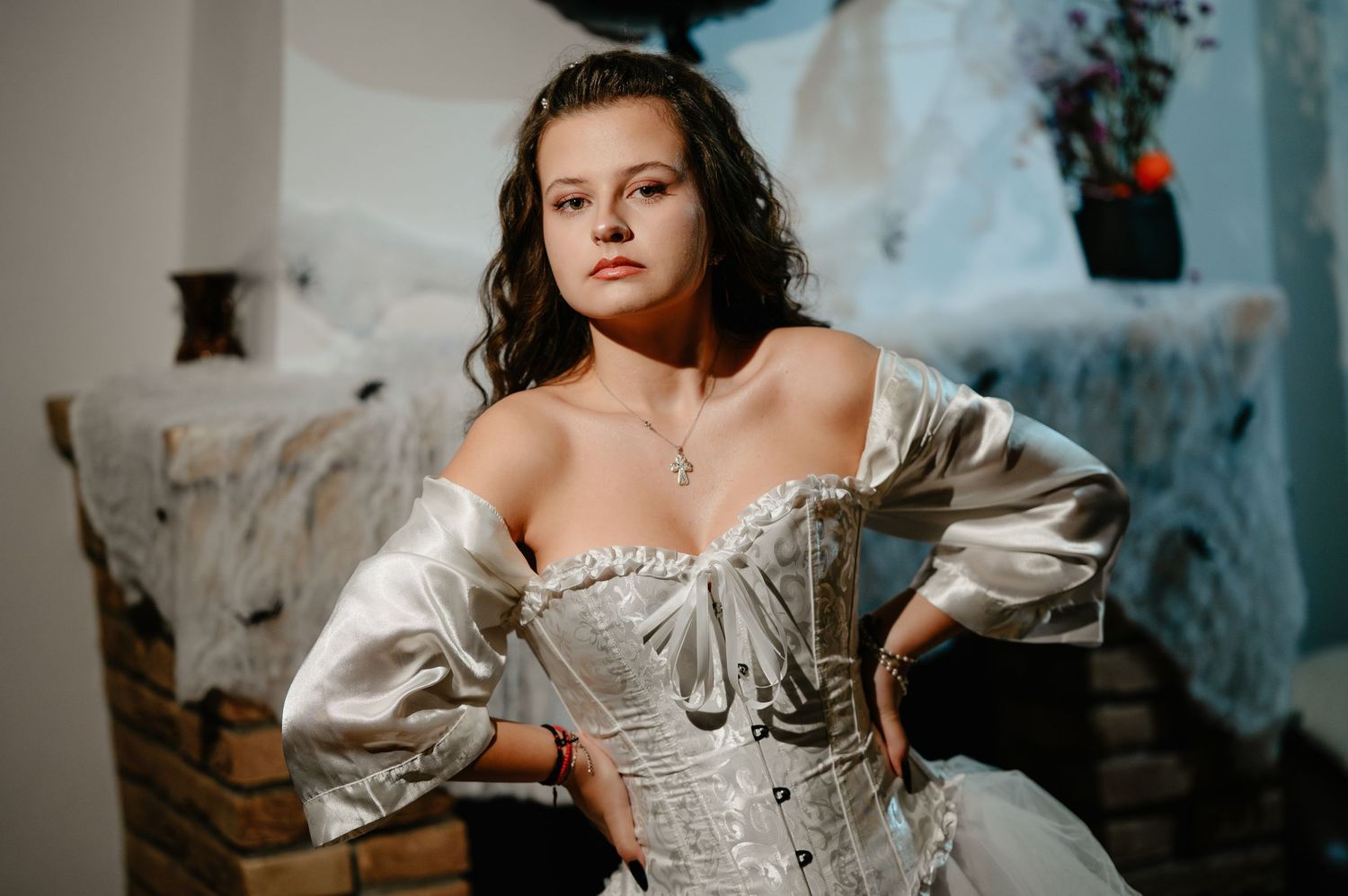 Young woman in elegant white corset gown with off-shoulder sleeves posing indoors at Halloween party.