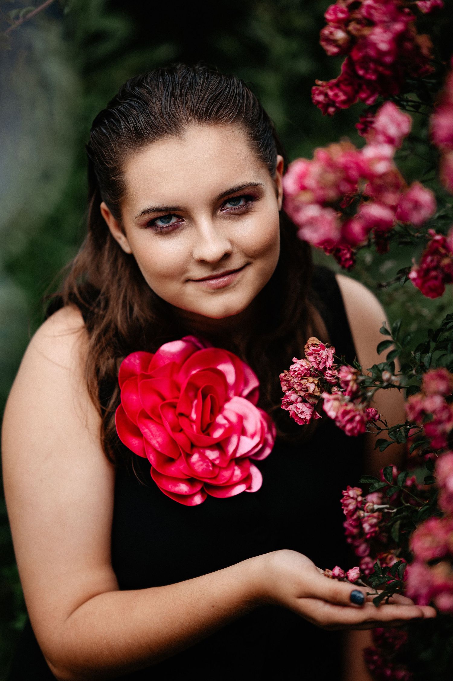 Young woman in black dress holds large red rose near blooming pink rose bush outdoors.