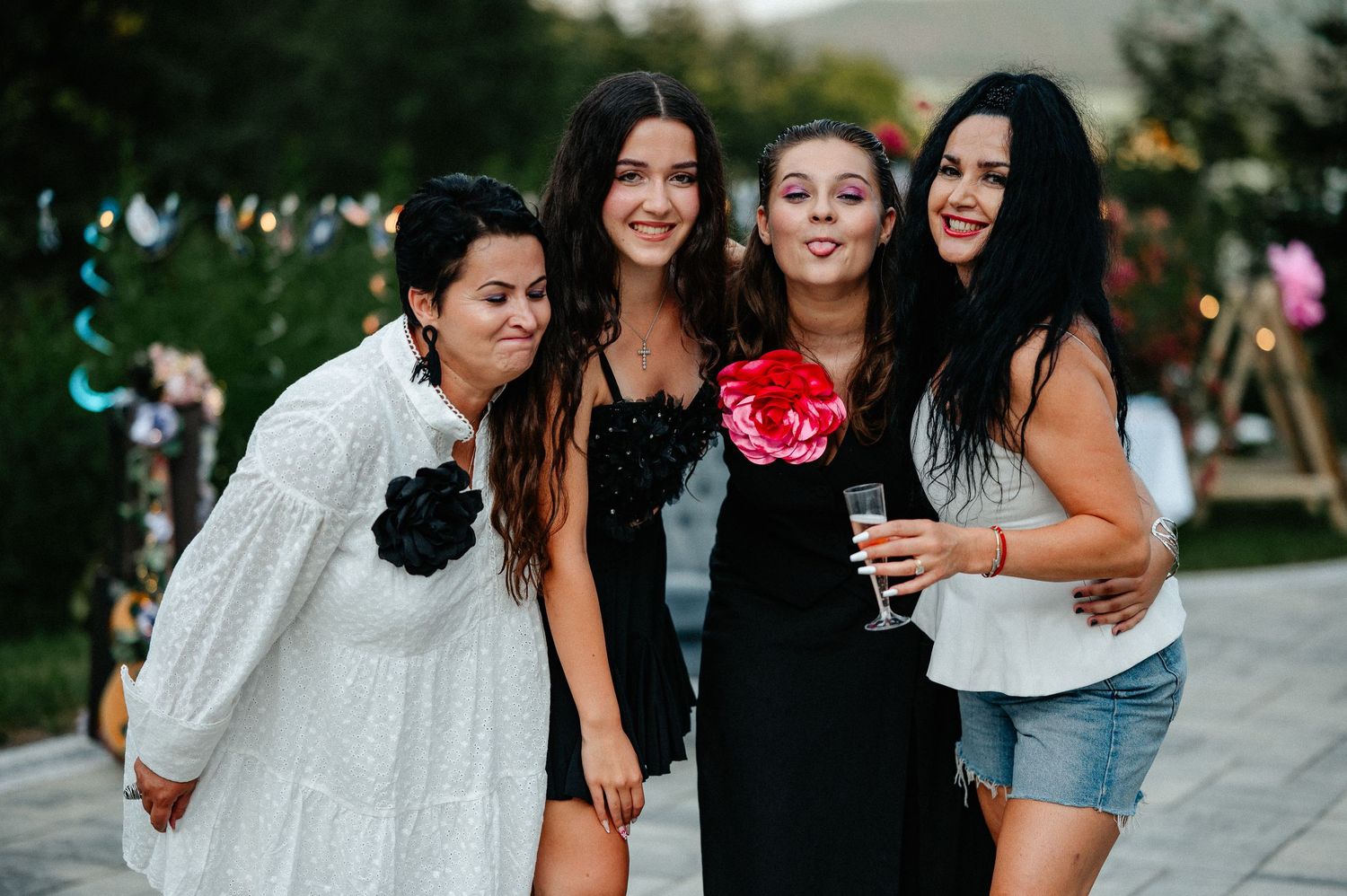 Four women posing together outdoors at an evening party, wearing elegant dresses with floral accessories.
