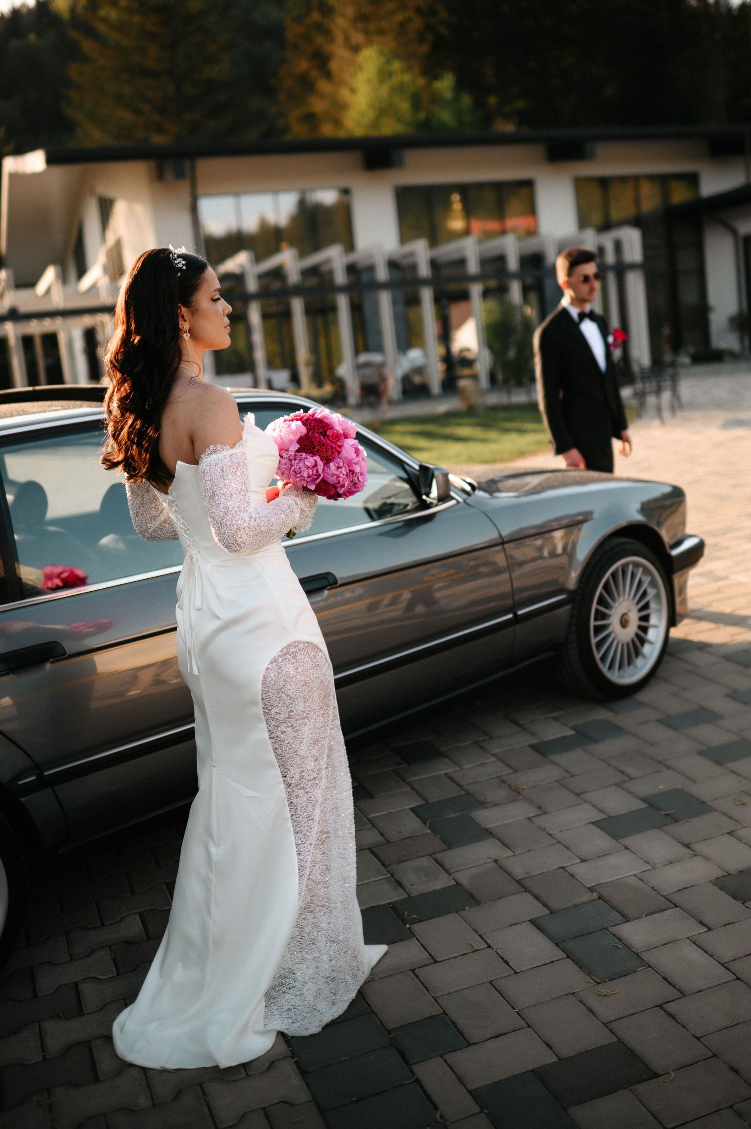 Elegant bride in lace off-shoulder gown with pink bouquet posing beside luxury car.