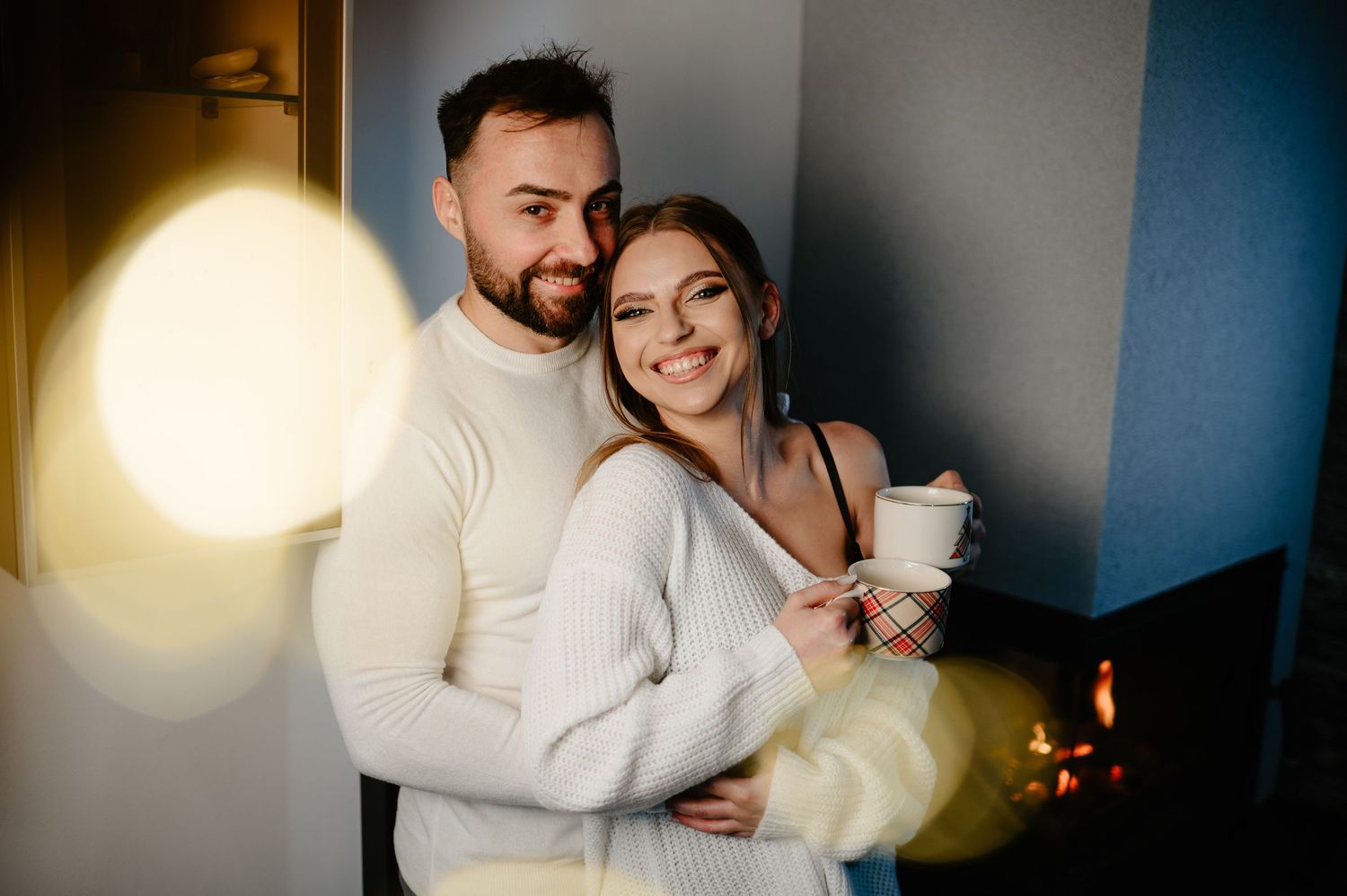 Happy couple in white sweaters sharing a warm embrace by firelight during Christmas.