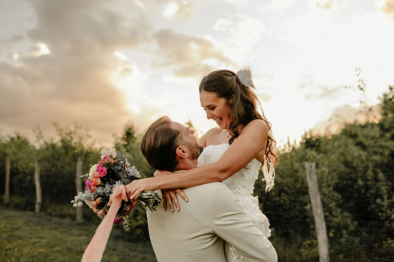 Groom lifting bride joyfully against dramatic sunset sky in vineyard wedding photo.