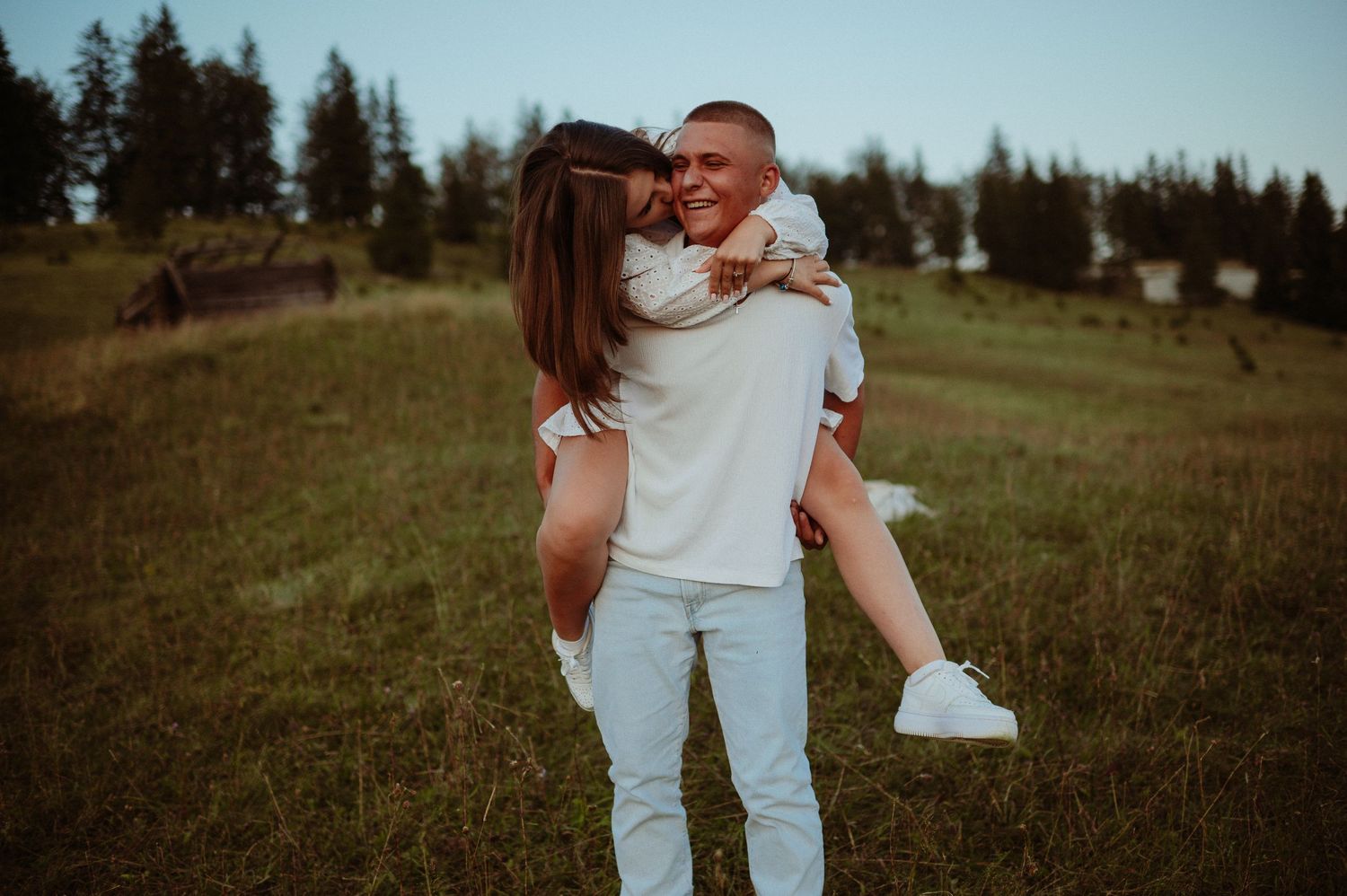 Smiling man carrying woman in white dress on green hillside with pine trees in background at golden hour.