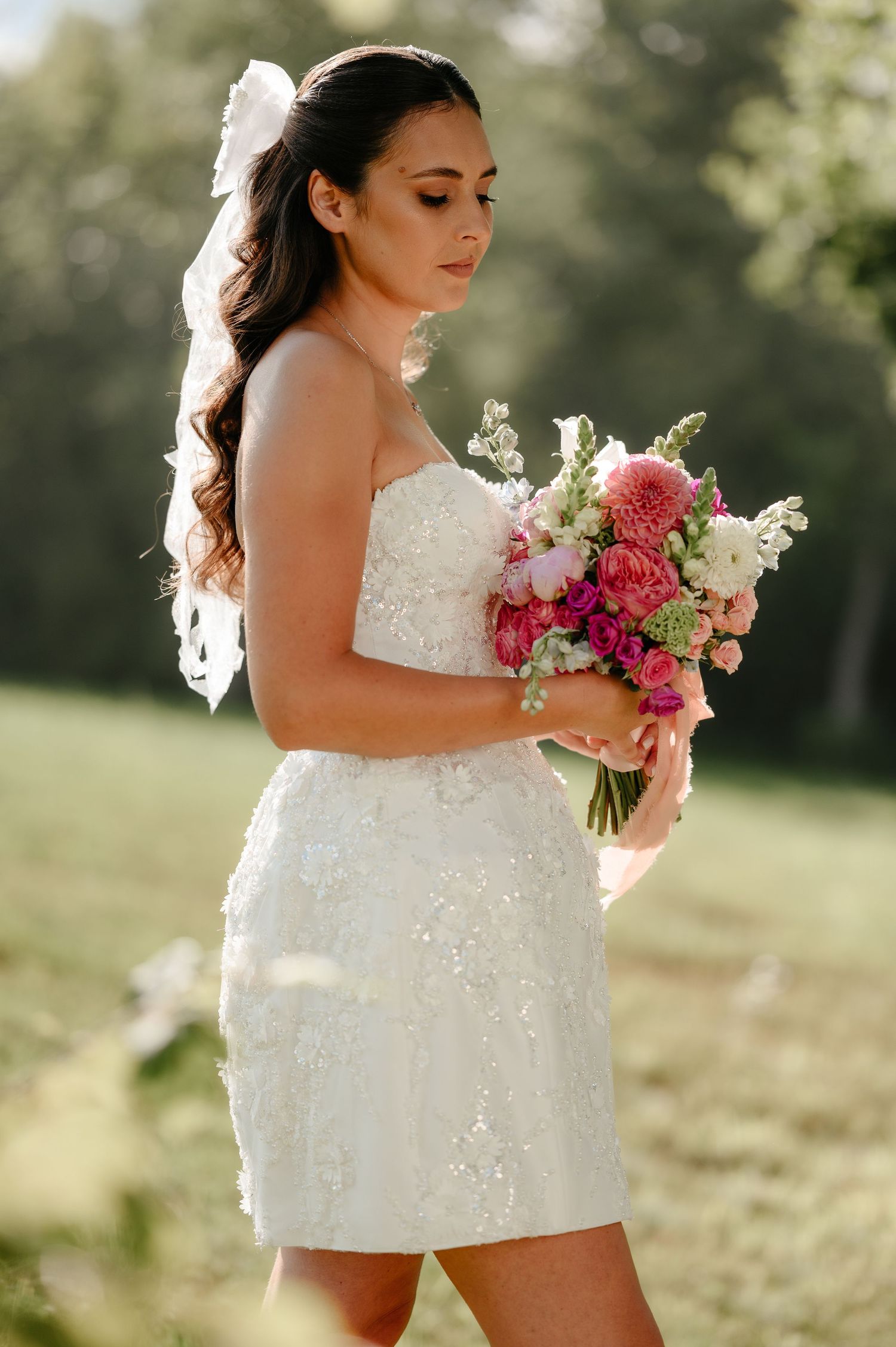 Elegant bride in white strapless gown holding a pink and white bouquet in a sunny garden.