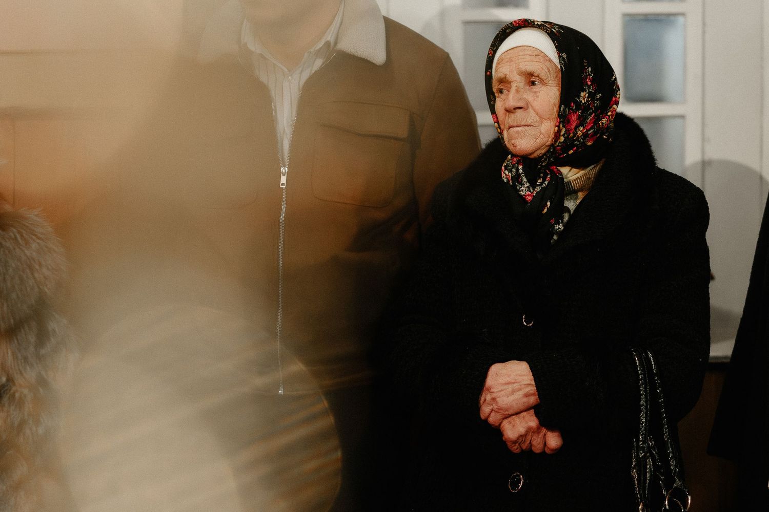 Elderly woman in black coat and floral headscarf standing solemnly at a religious ceremony.