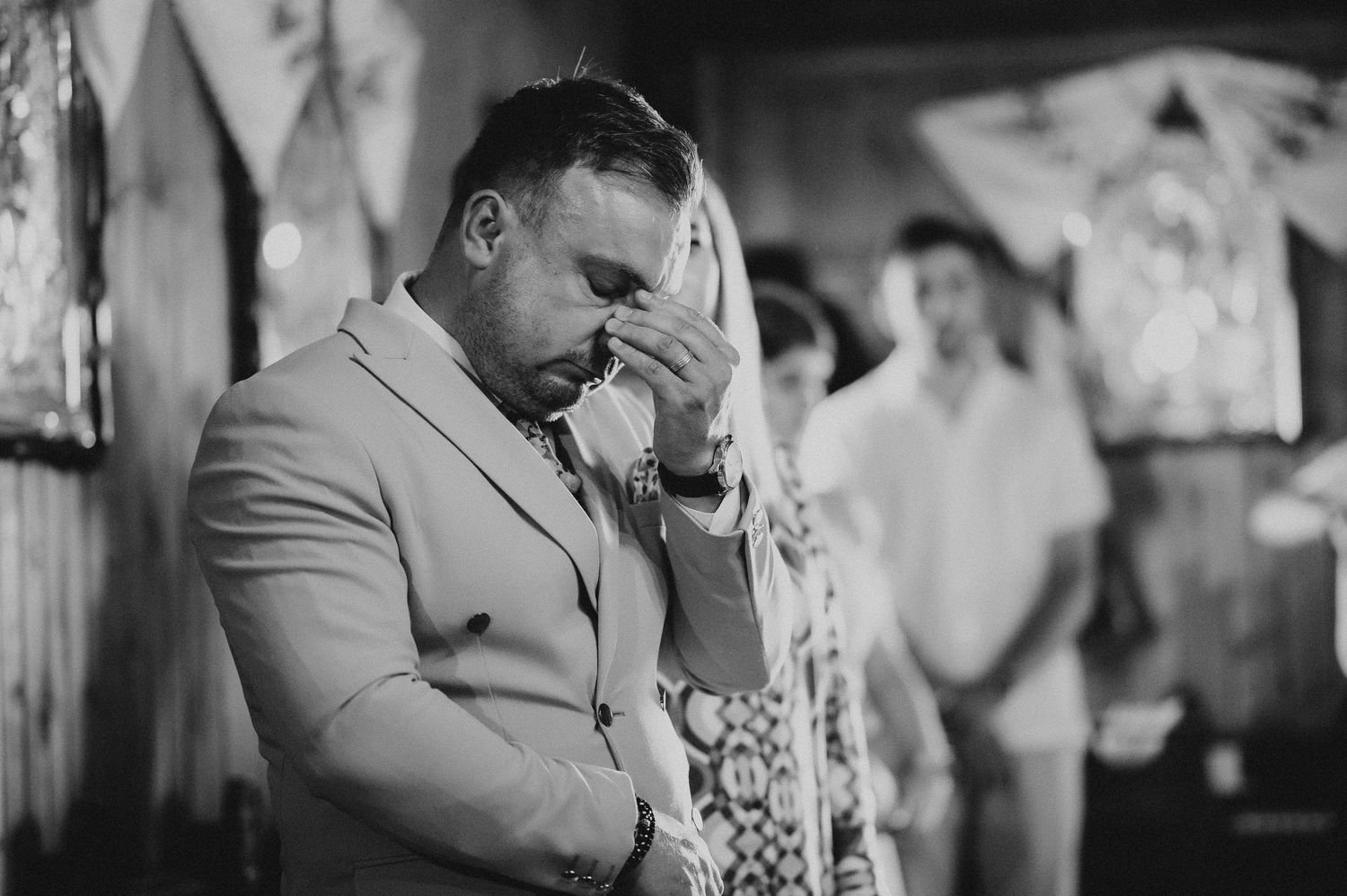 Emotional man in beige suit wipes tears during a heartfelt religious christening ceremony.