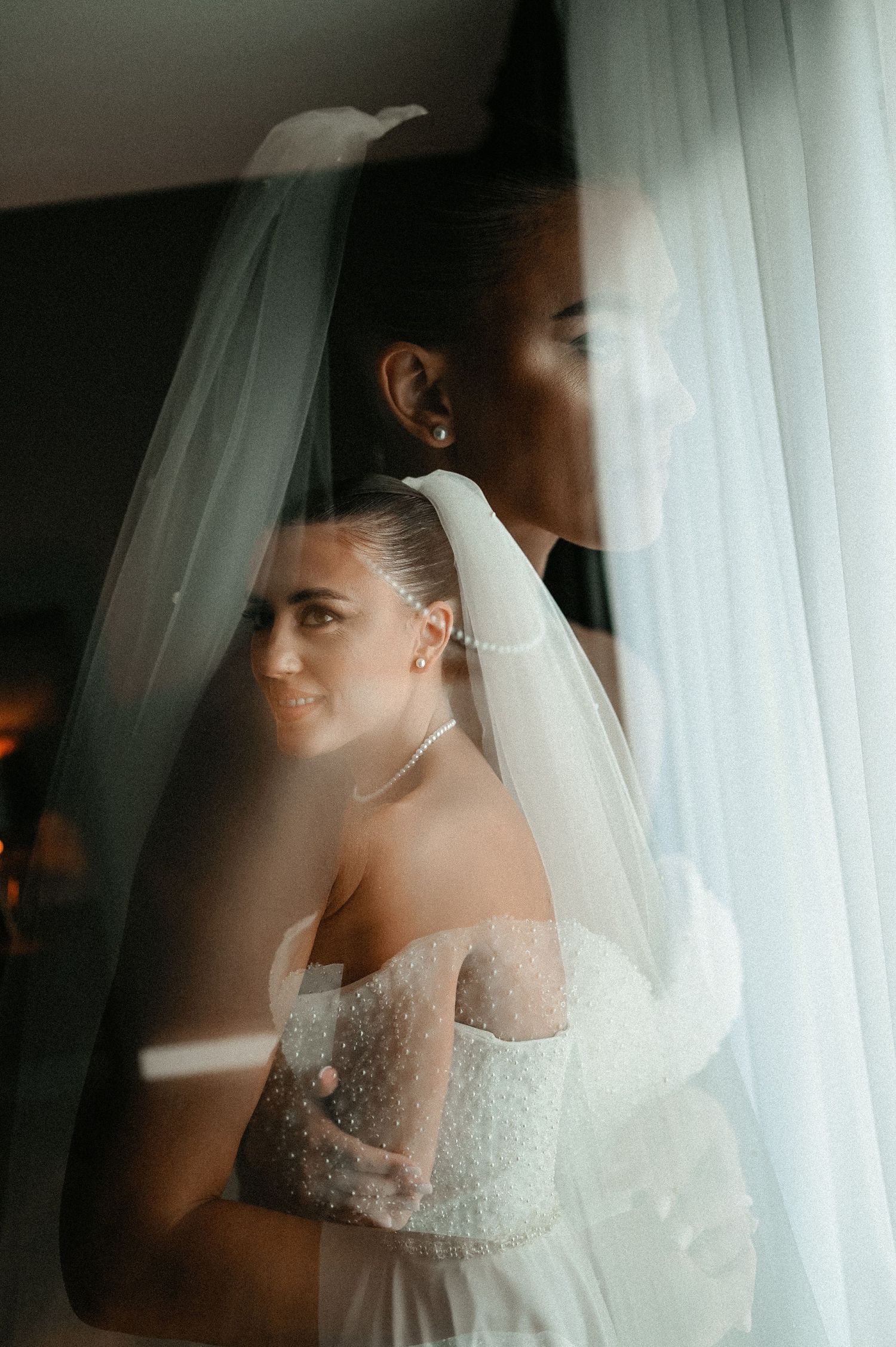 Bride smiling softly beneath a flowing veil in a dotted off-shoulder wedding dress by a window.