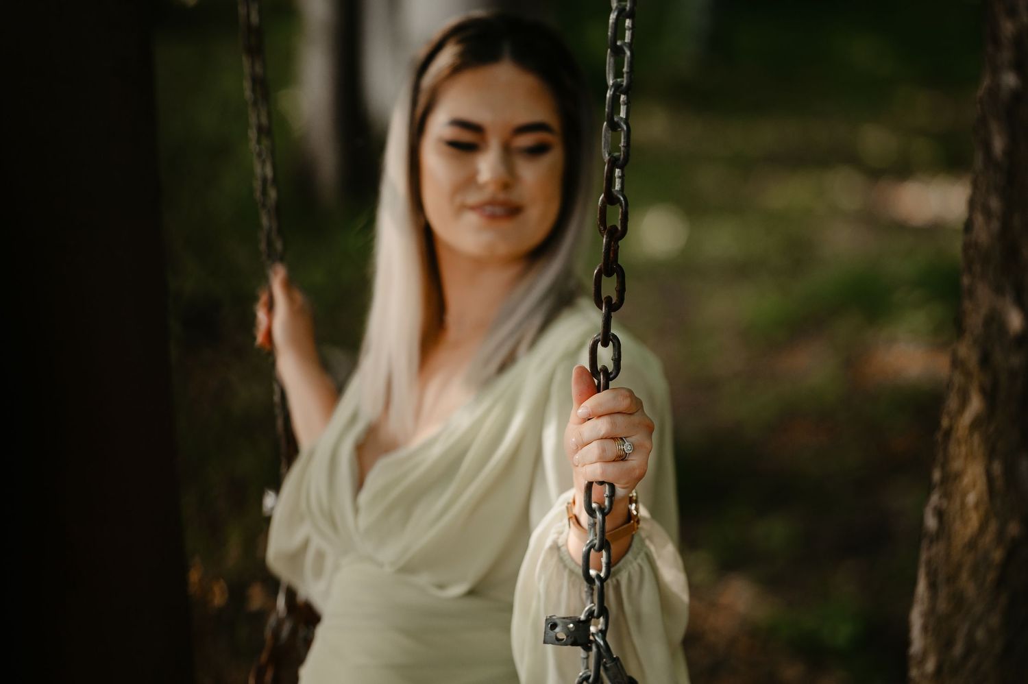 Young woman in sage green dress sitting on a chain swing in a blurred forest background.