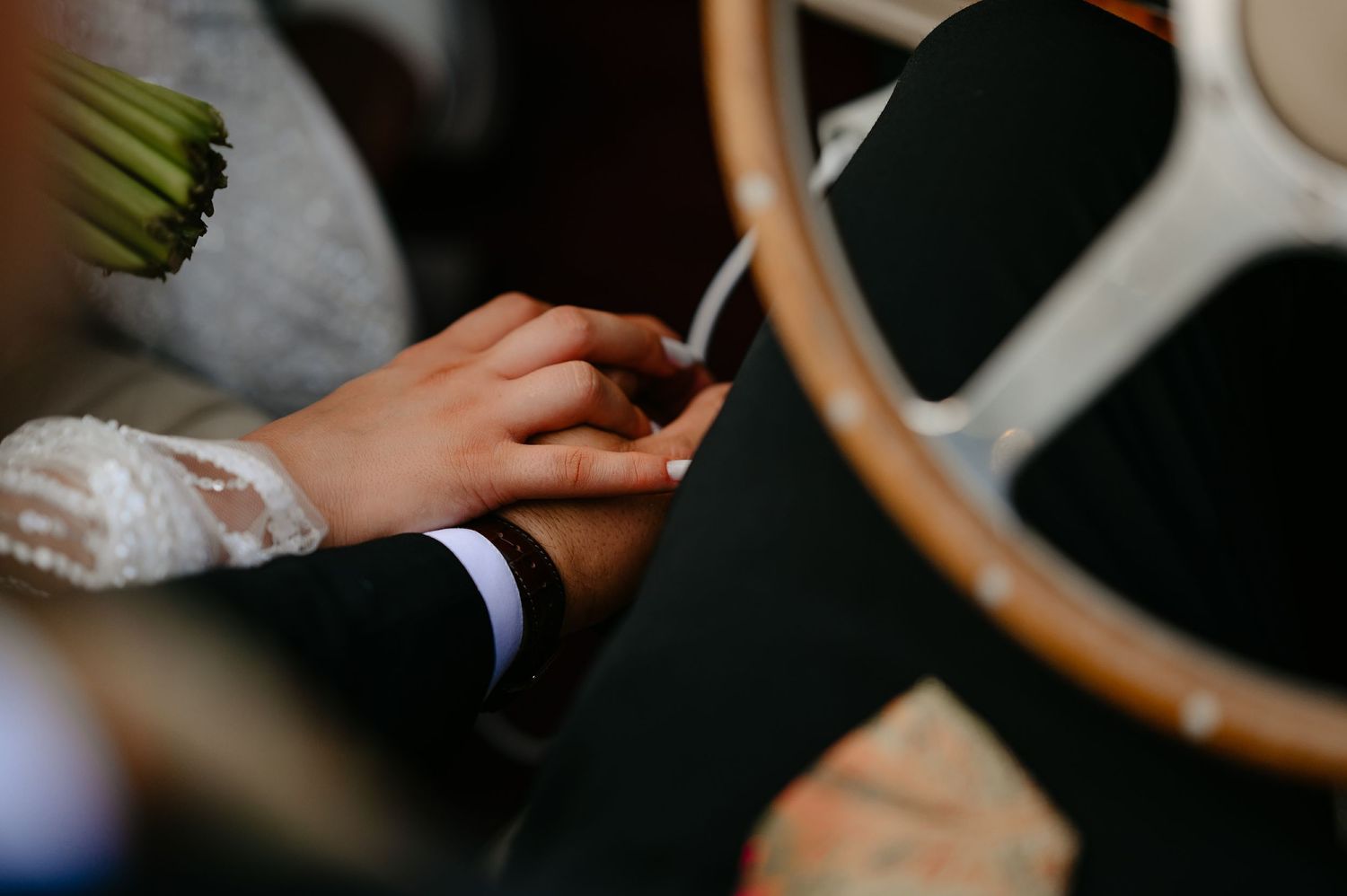 Close-up of bride and groom holding hands near a vintage car steering wheel on wedding day.