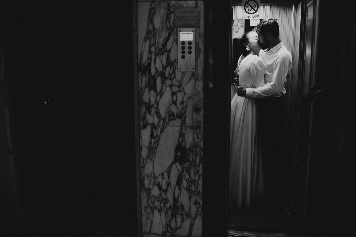 Black and white artistic photo of couple kissing in dimly lit elevator doorway in wedding attire.