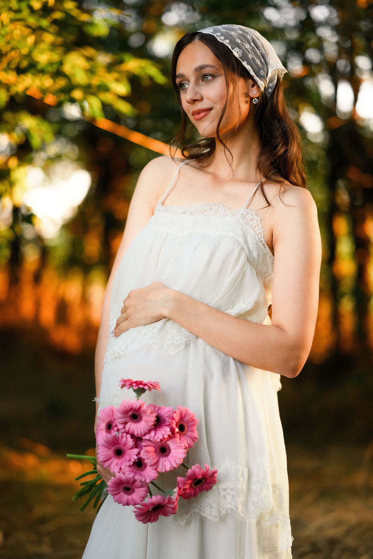 Pregnant woman in white dress holding pink gerbera daisies, standing outdoors in golden sunset light.