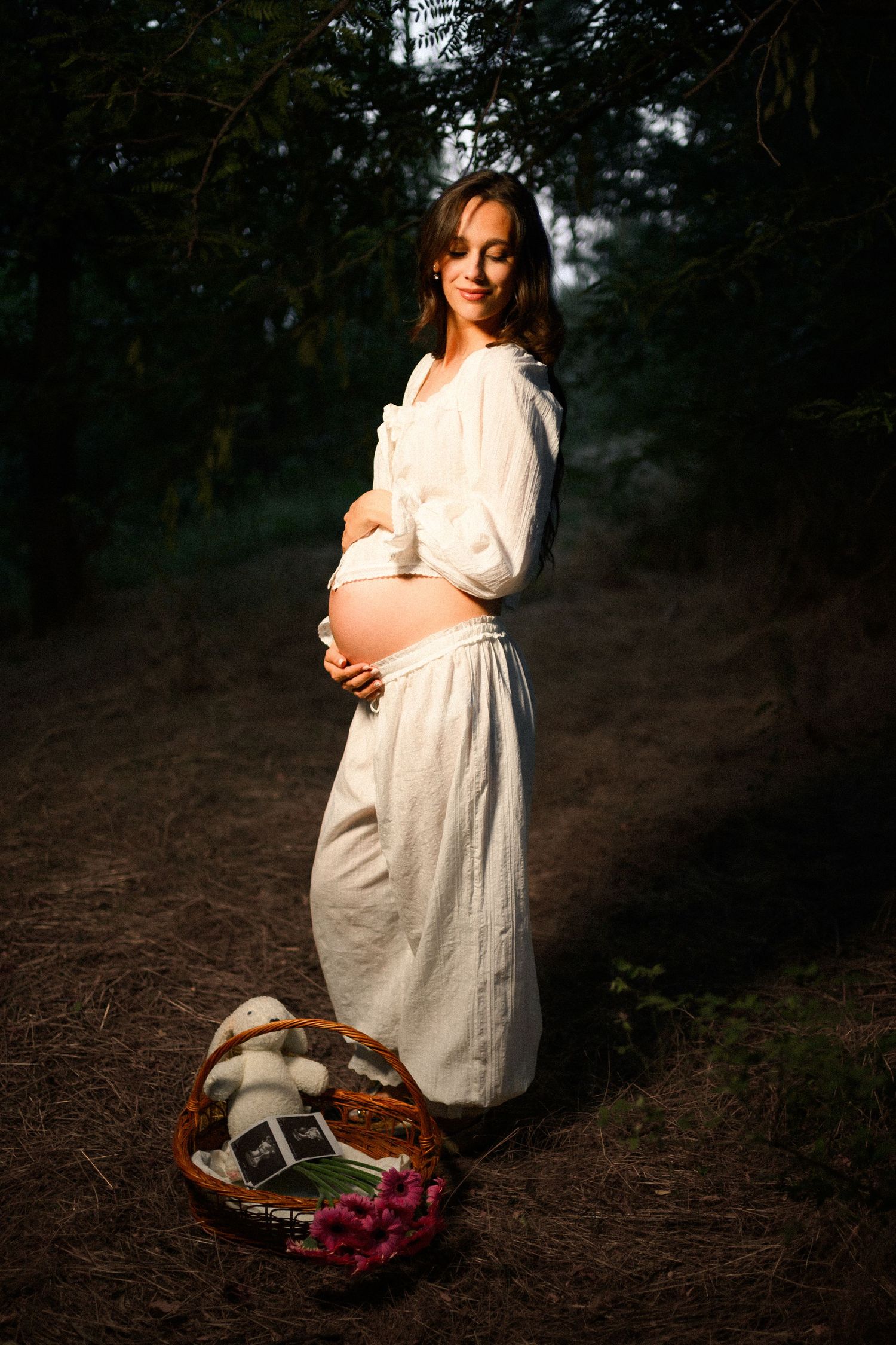 Pregnant woman in white dress holding belly outdoors in forest, with basket of ultrasound photos and flowers nearby.