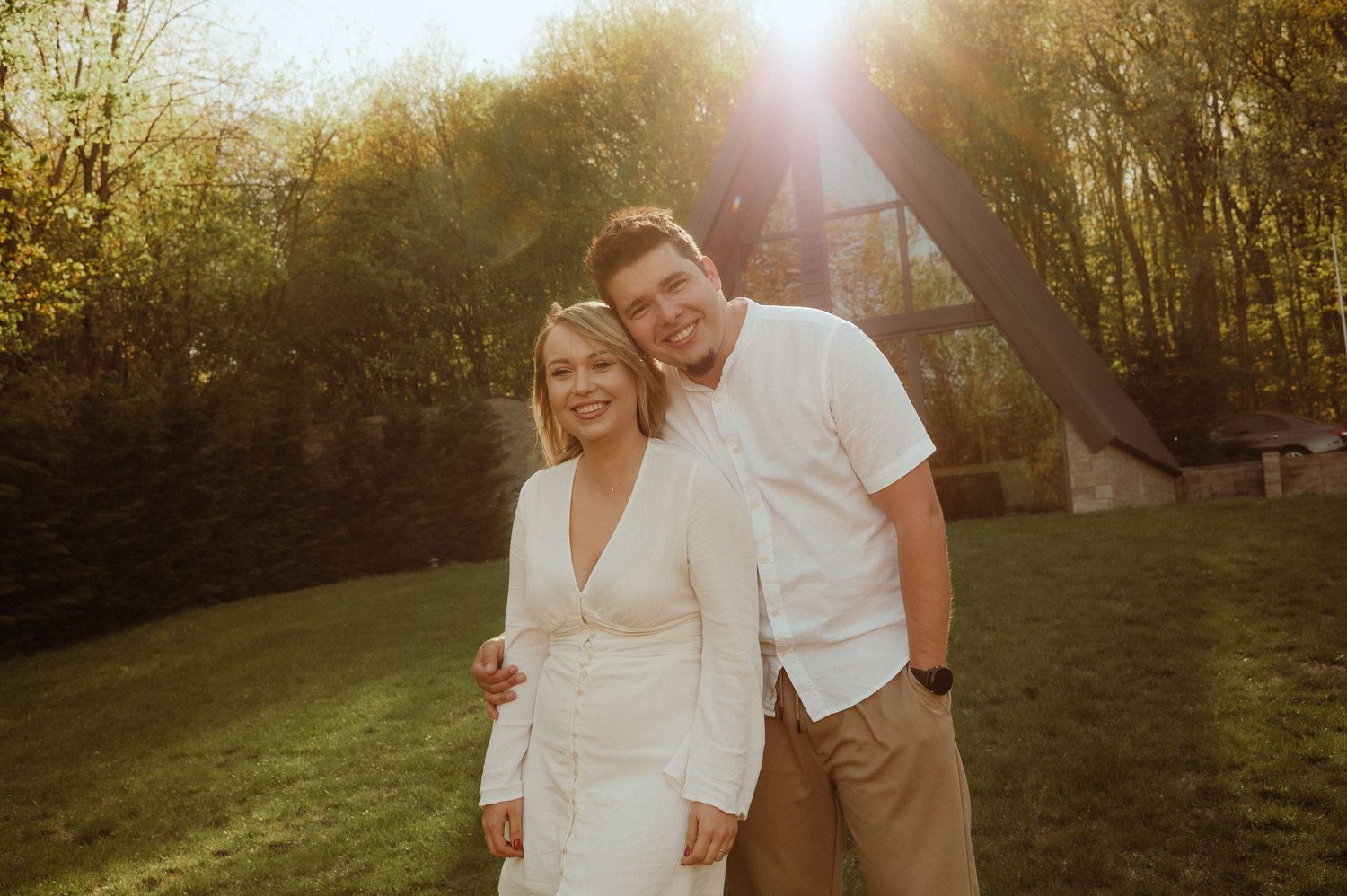 Smiling couple embracing outdoors in warm sunset light with trees and A-frame structure behind them.