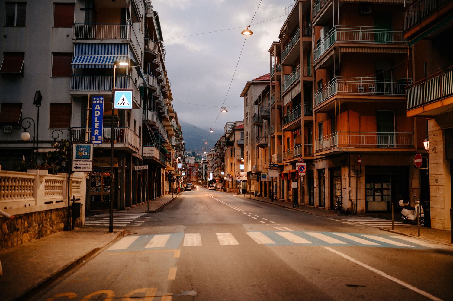 Empty European street at dusk with buildings, streetlights, and a pedestrian crosswalk.