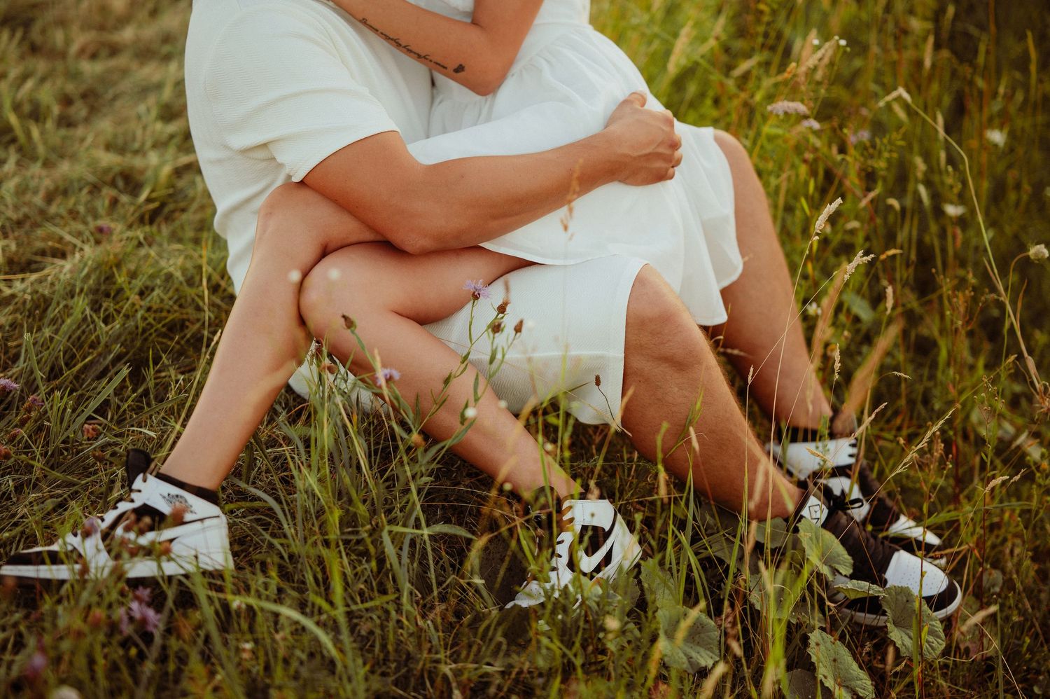 Couple sitting together in tall grass wearing matching black and white sneakers outdoors.