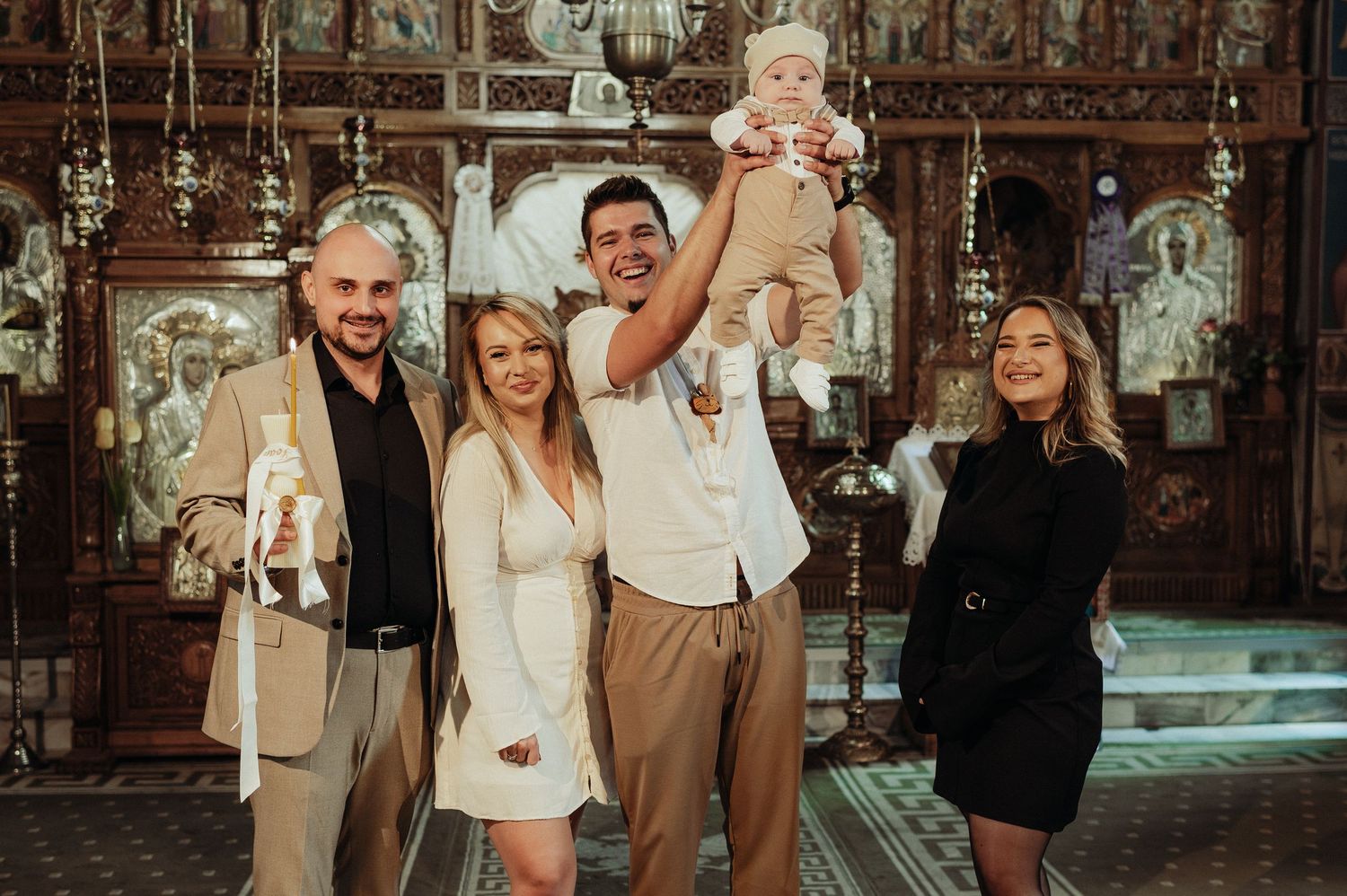 Joyful family lifting baby in the air inside an ornate Orthodox church after a baptism ceremony.