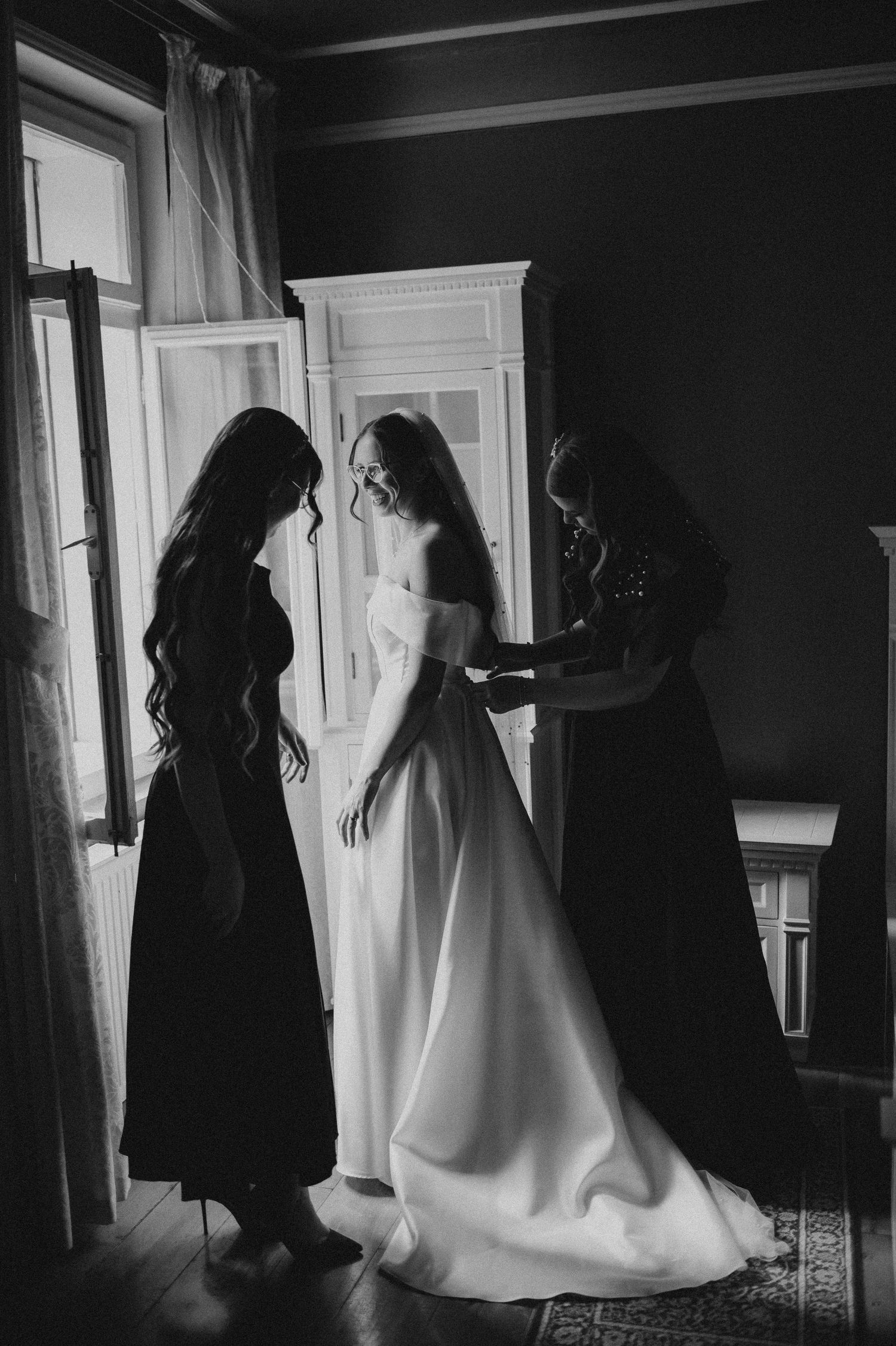 Bridesmaids helping bride into white wedding dress by window in black and white.