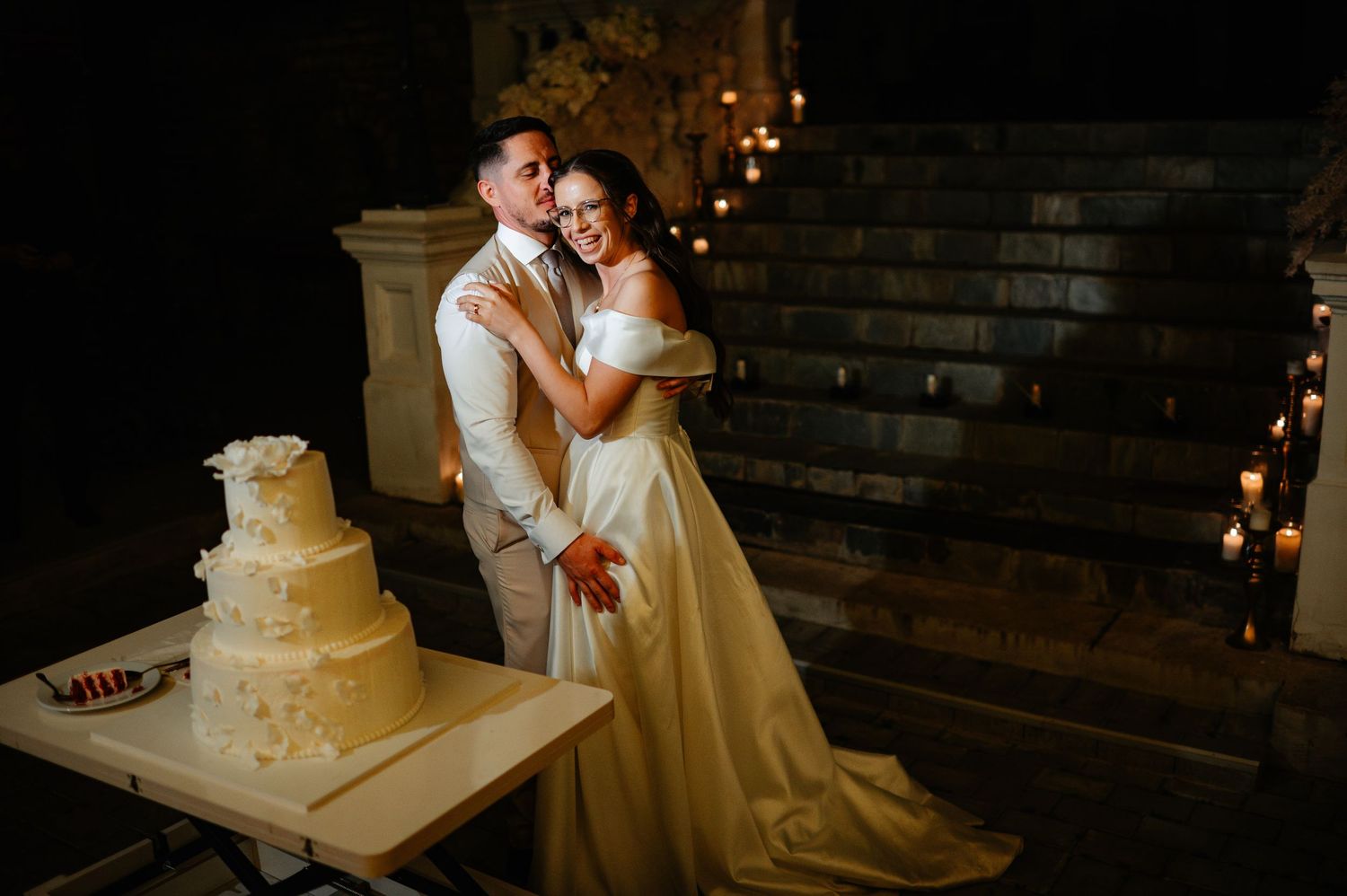 Bride and groom pose lovingly beside elegant tiered wedding cake on candlelit staircase at night.