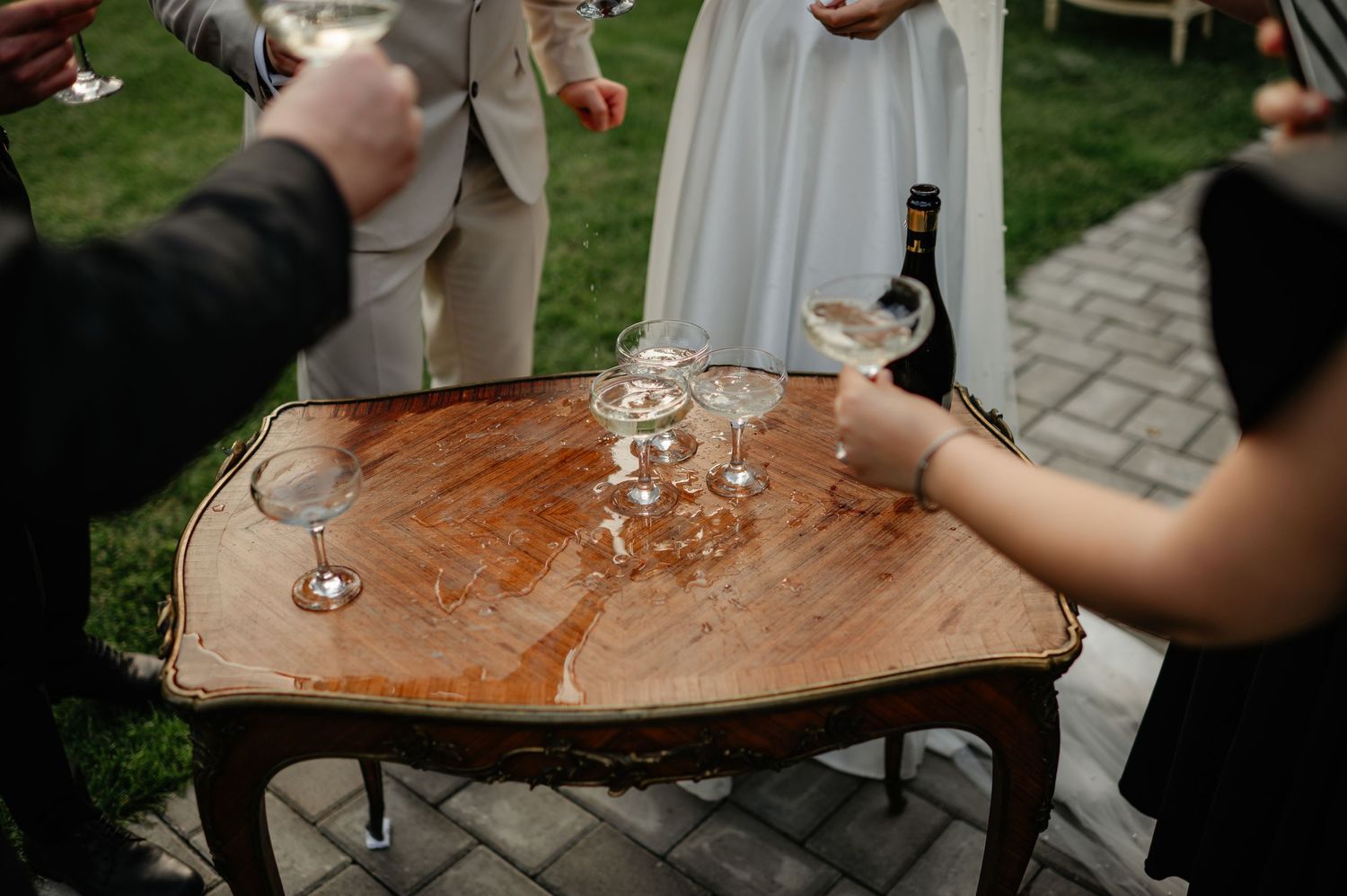 Guests clinking champagne coupe glasses on a rustic wooden table at an outdoor wedding reception.
