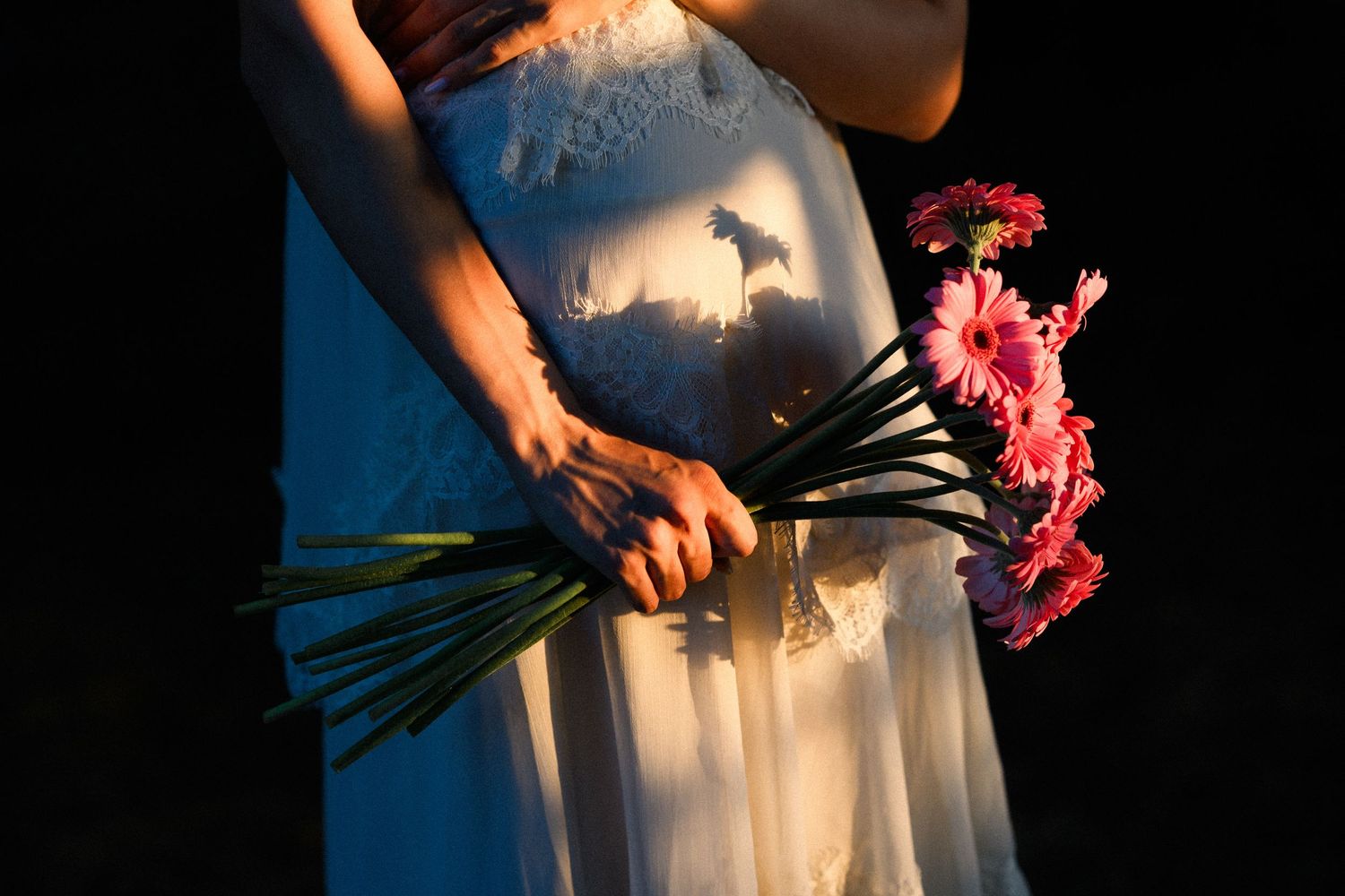 Bride in white lace gown holding pink gerbera daisy bouquet with dramatic sunlight and dark background.