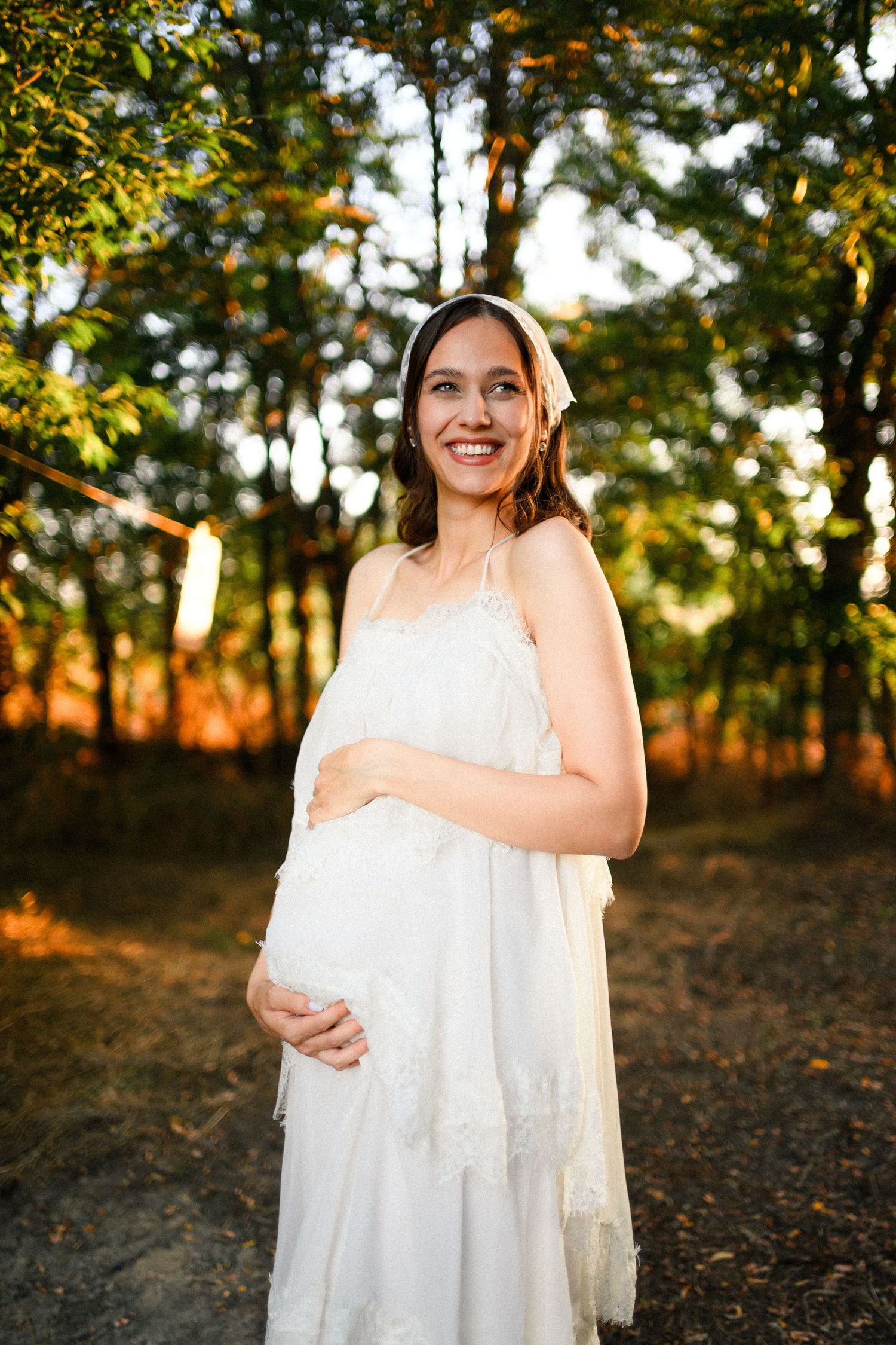 Smiling pregnant woman in white dress holding belly outdoors in sunlit forest during golden hour.
